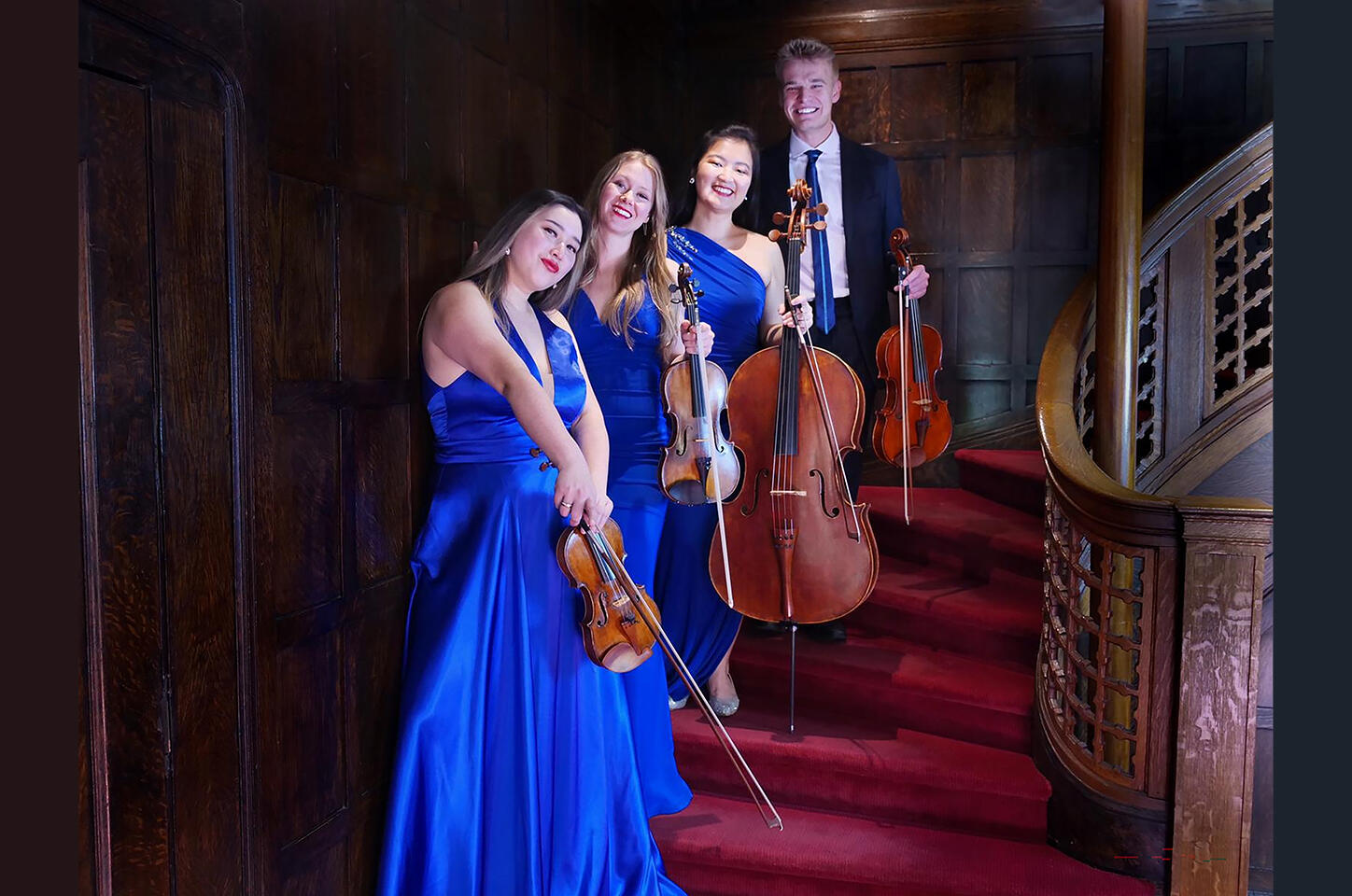 Photo of the members of the Ulysses Quartet standing on a staircase holding instruments. Three woman (2 Asian, 1 White) wear blue gowns. 1 white man wears a suit.