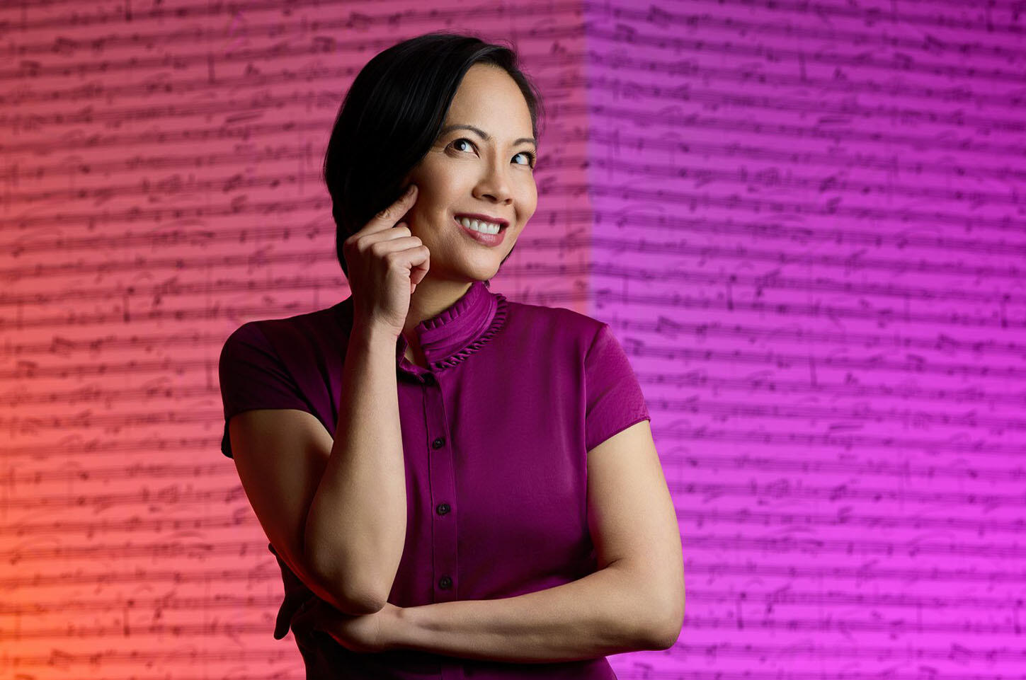 Photo of smiling Vivian Fung (Asian woman) wearing plum colored blouse standing in front of a purple and pink wall covered with musical notation.