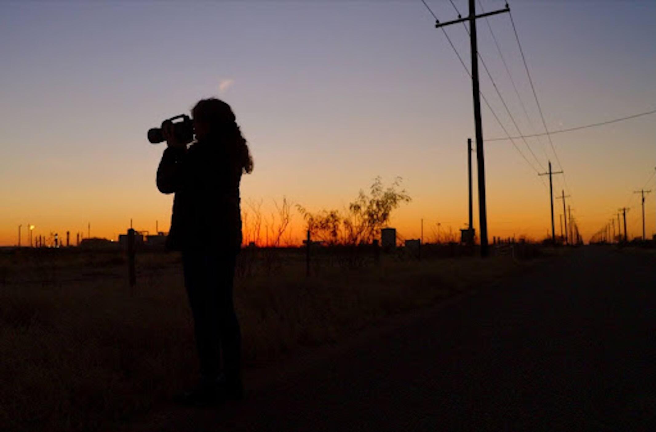 Sillhouette of a person holding a camera at dusk. The sun is setting in the background.