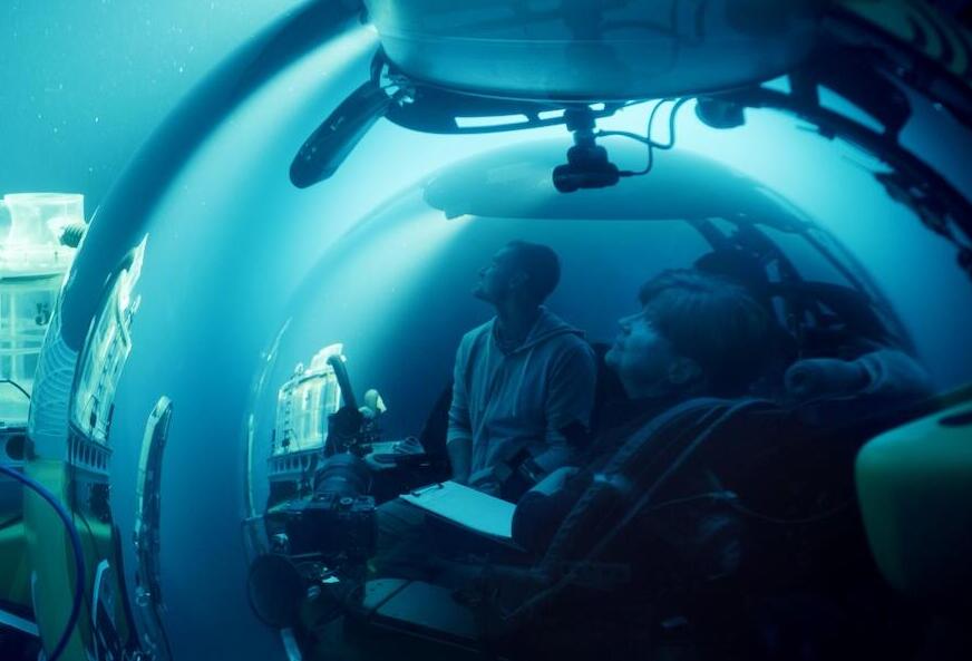 Two people in a glass-dome underwater submersible surrounded by blue ocean water. 