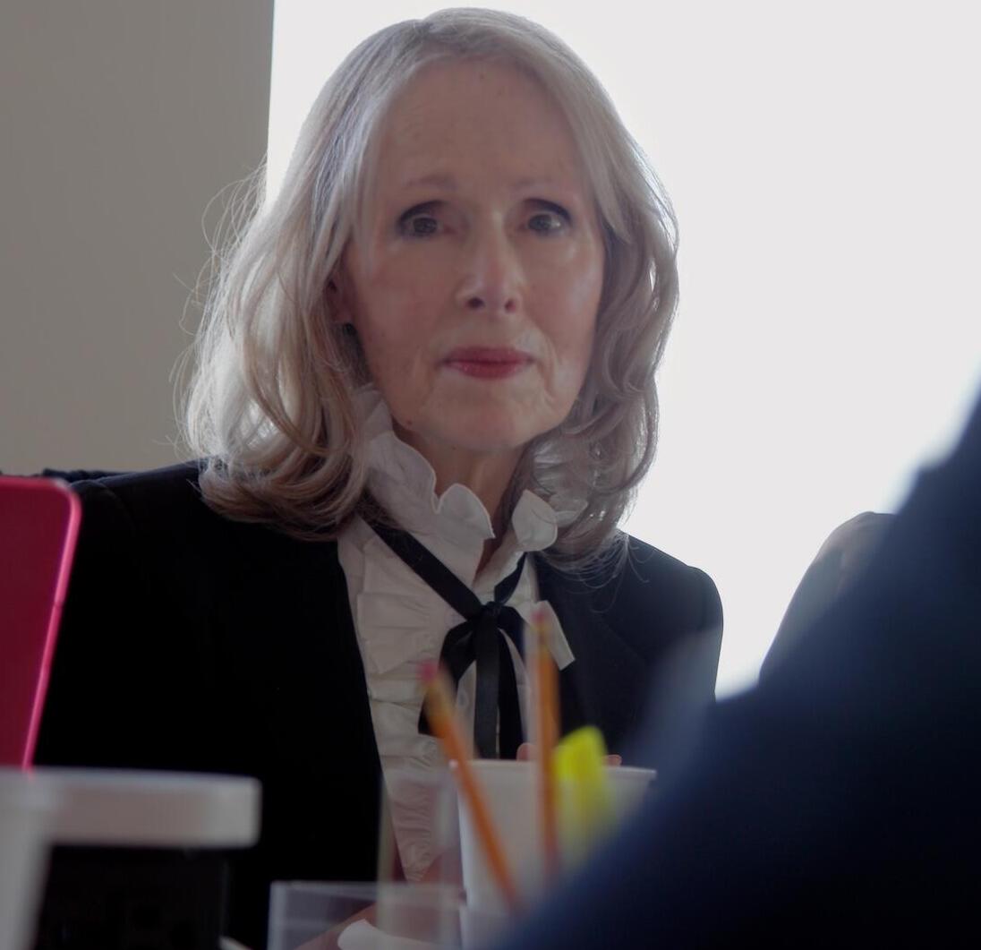 Silver-haired, professionally dressed woman seated at a table looking at the camera.