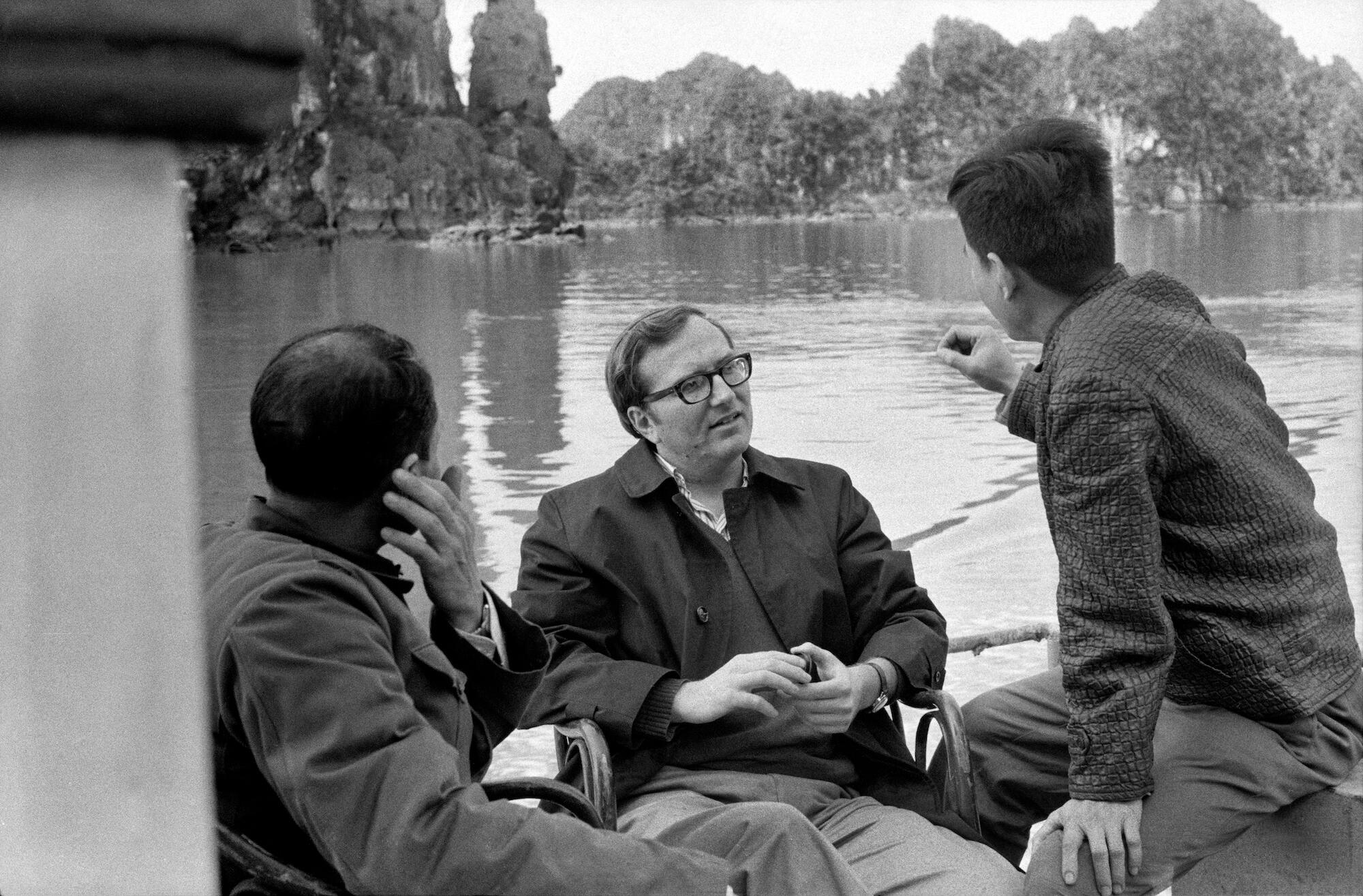Older black and white photo of three men talking. A pond and trees are visible in the background..
