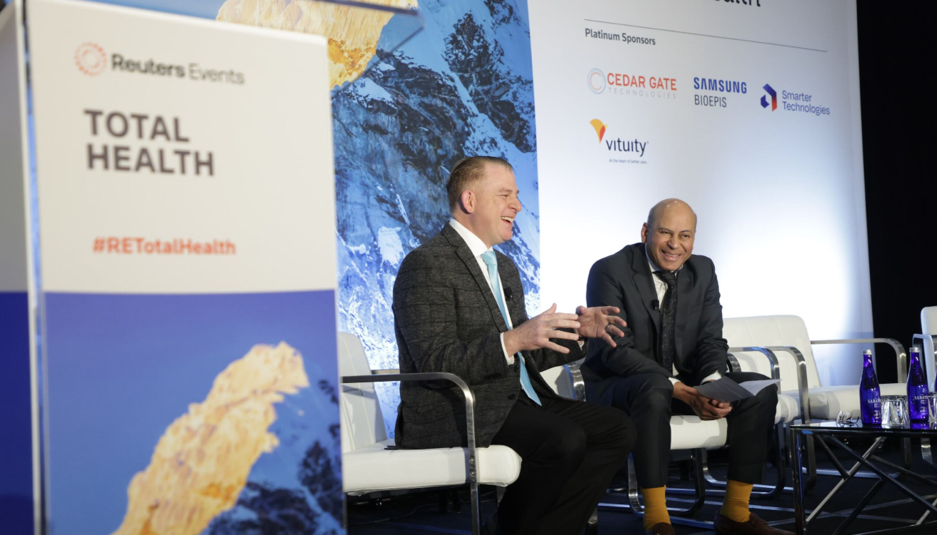 Two male speakers laughing and in conversation on stage at the Reuters Events Total Health conference, with platinum sponsor logos including Cedar Gate Technologies, Samsung Bioepis, Smarter Technologies