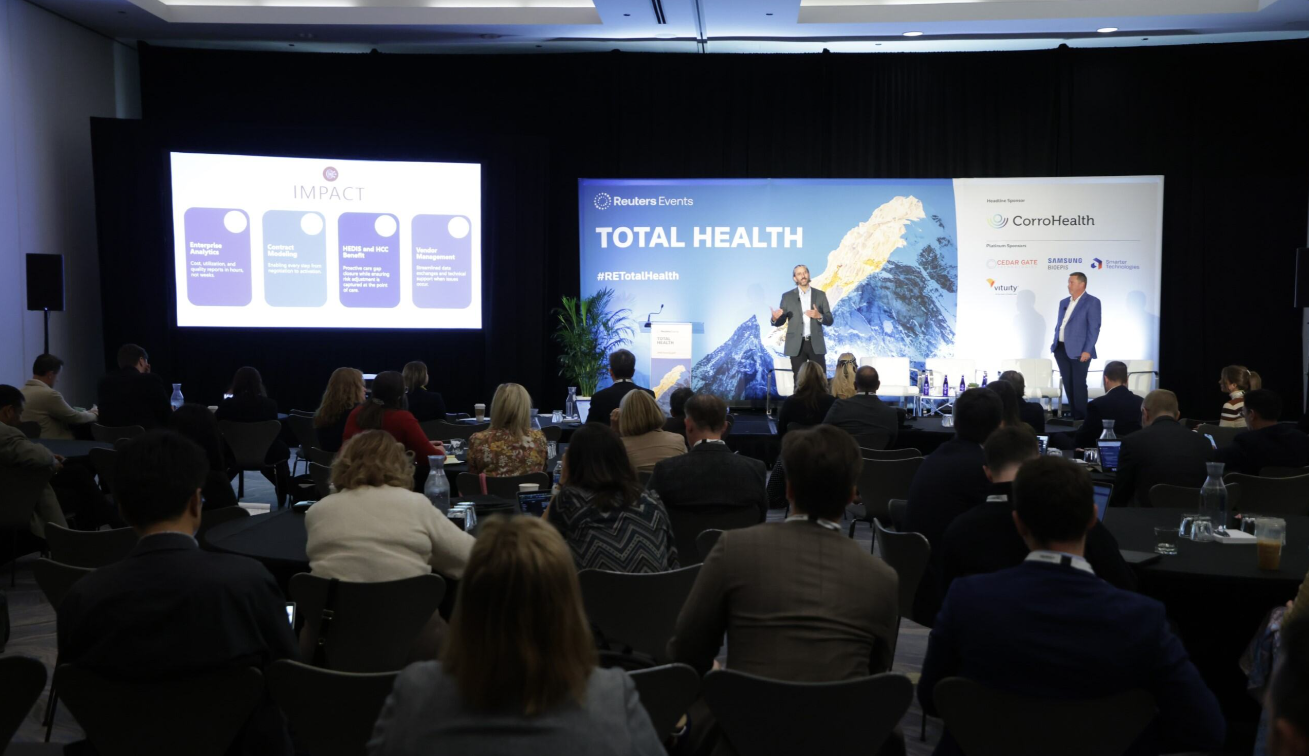 A speaker presenting an "Impact" slide to a seated audience at the Reuters Events Total Health conference, with sponsor logos for CorroHealth, Cedar Gate, Samsung Bioepis, Smarter Technologies, and Vituity visible on the backdrop.