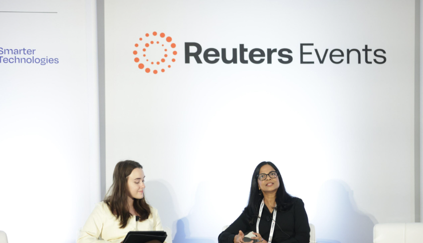 Two women seated on a stage in conversation at a Reuters Events conference — a young woman in a cream blazer holding a tablet on the left, and a woman in a black blazer wearing glasses and a lanyard on the right — with the Reuters Events logo and Smarter Technologies sponsor banner visible in the background.