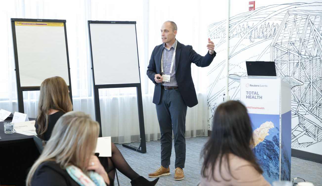 A speaker presenting to a seated audience at the Reuters Events Total Health conference, with whiteboards and a branded podium visible in the background.