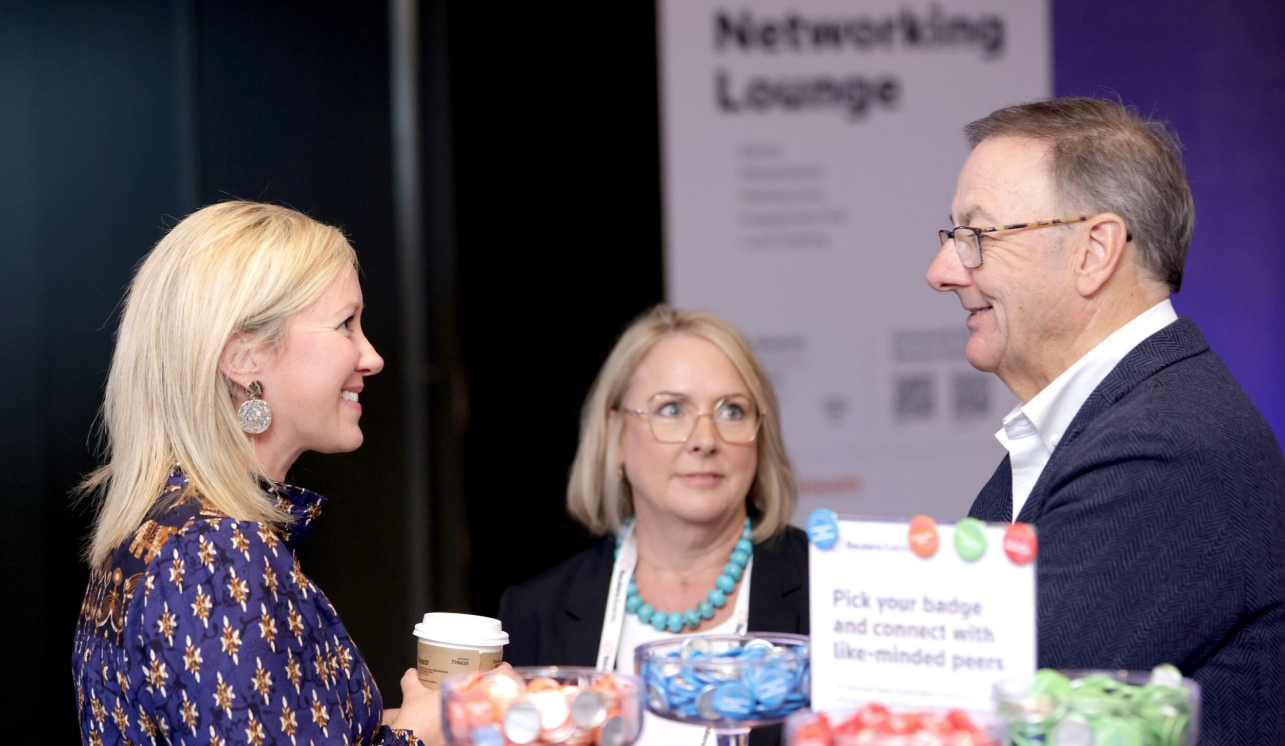 Three attendees networking in the Networking Lounge at the Total Health conference — a blonde woman in a navy floral dress holding a coffee cup, a woman in a black blazer with a turquoise necklace, and a man in a dark blazer with glasses — engaged in conversation beside a table with coloured badges and a sign reading "Pick your badge and connect with like-minded peers."