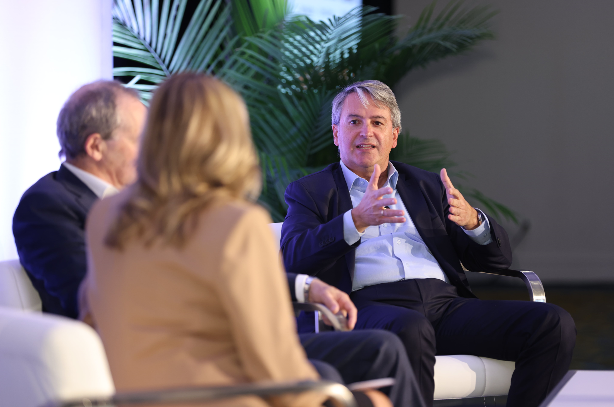 Fireside chat at Reuters Events: Automotive USA, male speaker in conversation on stage, gesturing while discussing industry topics, with two panelists in the foreground and plants as backdrop.