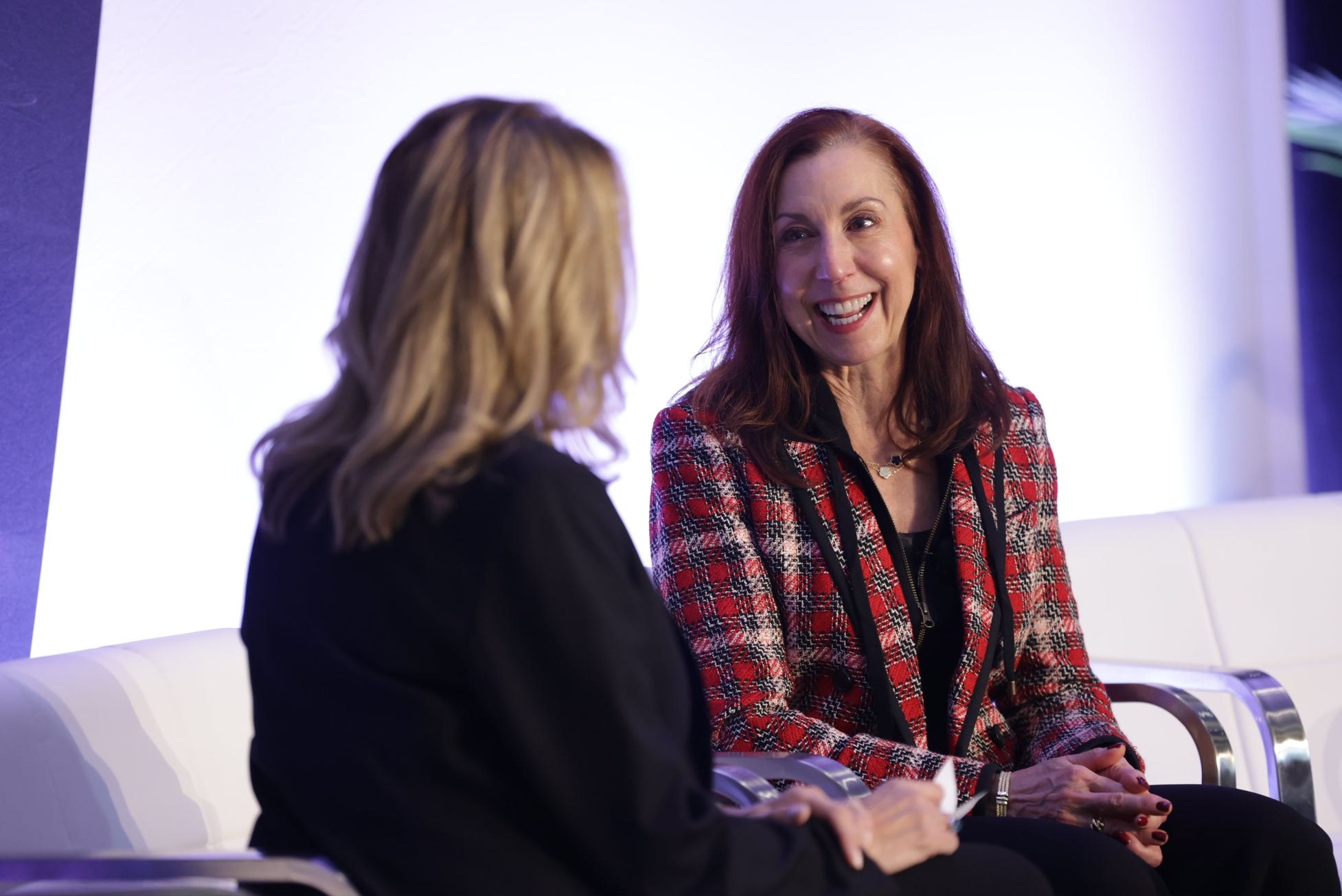 Fireside chat at Reuters Events: Automotive USA, two women speakers seated on a white stage sofa, engaged in discussion and smiling.