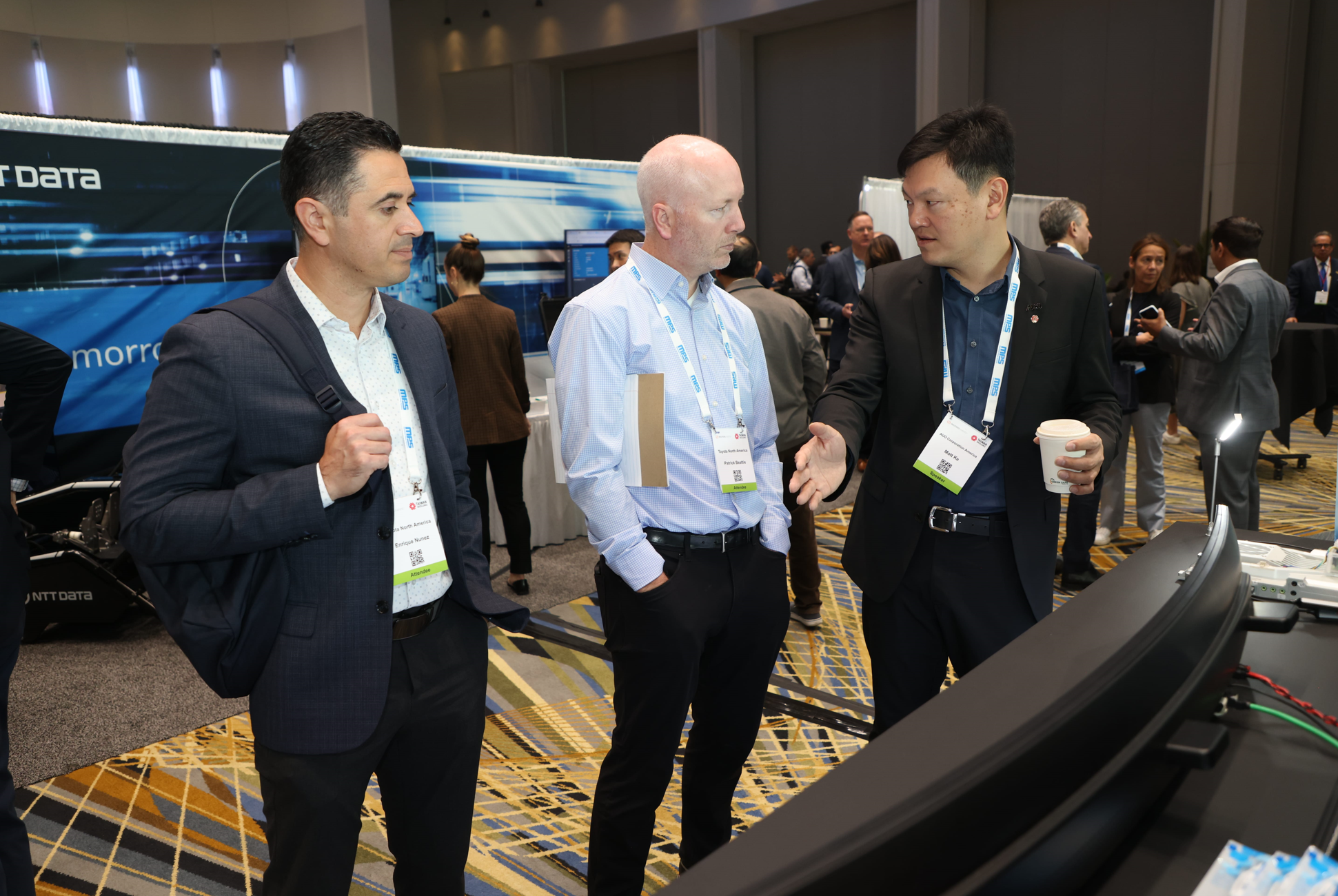Networking around a technology demo at Reuters Events: Automotive USA, three professionals in conversation at an exhibitor booth, with curved display equipment and event badges visible.