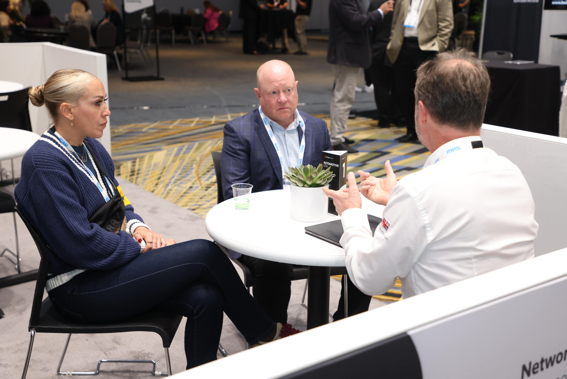 Networking meeting at Reuters Events: Automotive USA, three professionals in conversation at a small round table on the expo floor, with event badges and materials visible.