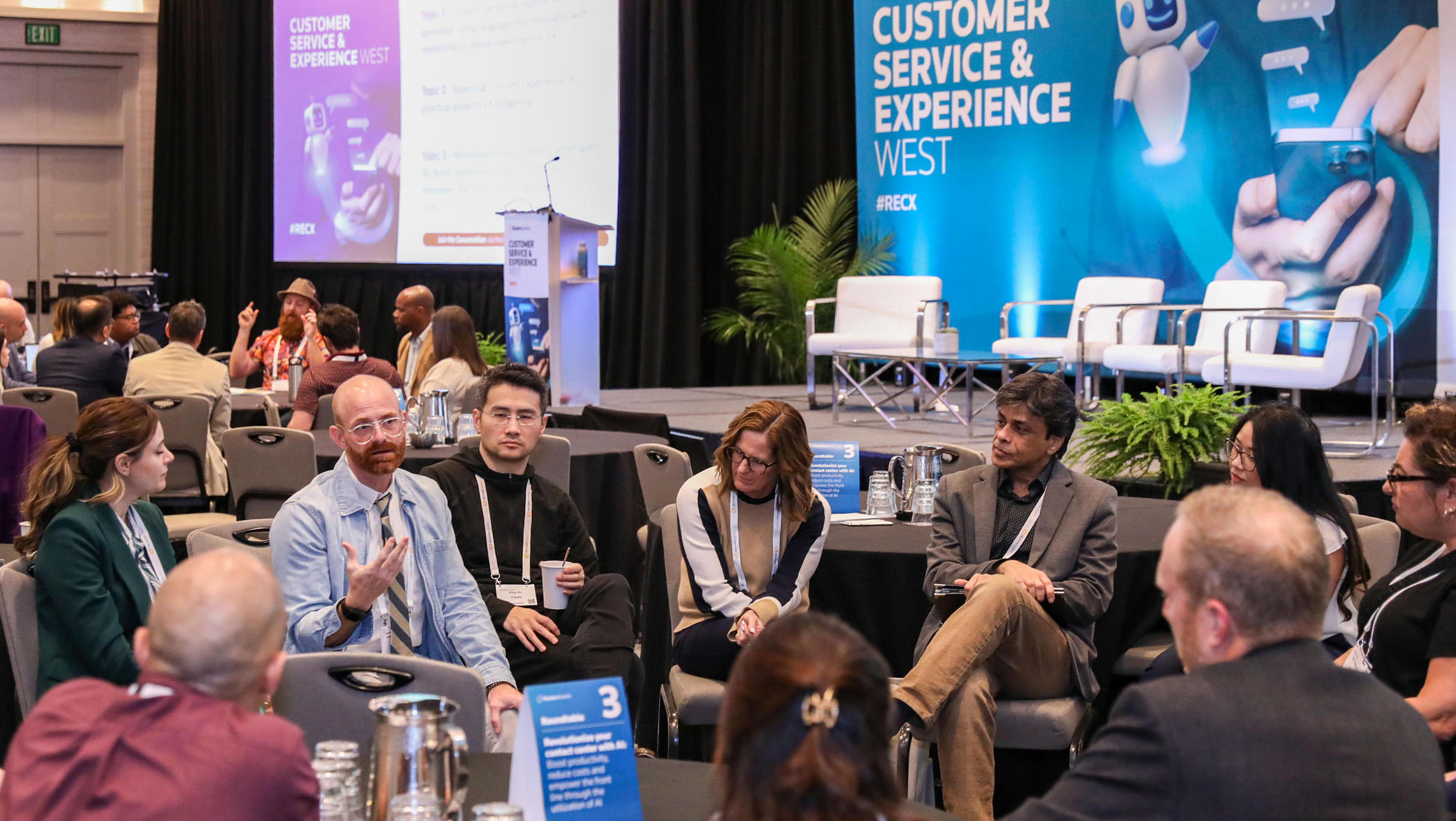 Customer Service & Experience conference small‑group roundtable: attendees discuss near stage with event branding and chairs in background