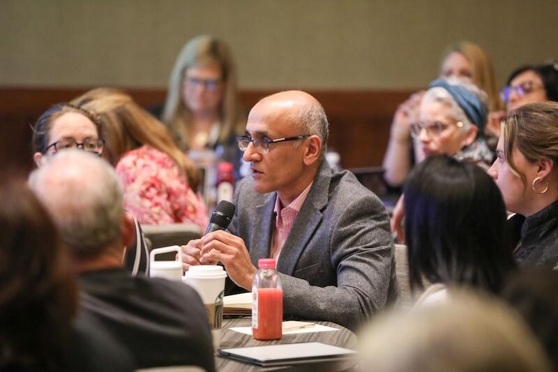 A person with a microphone talking to a group of people at a round table