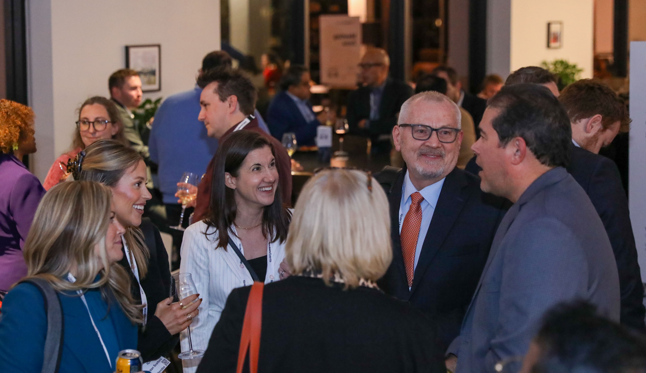 A crowd of professionally dressed attendees mingle at a Reuters Events networking reception, holding drinks and wearing lanyards, in a warmly lit indoor venue.