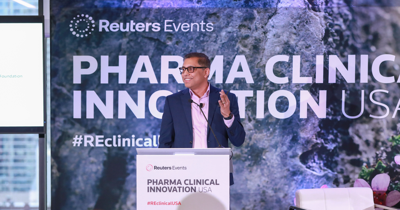 A man in a navy blazer and pink shirt speaks at a Reuters Events "Pharma Clinical Innovation USA" podium, gesturing with both hands, with a large branded backdrop behind him.