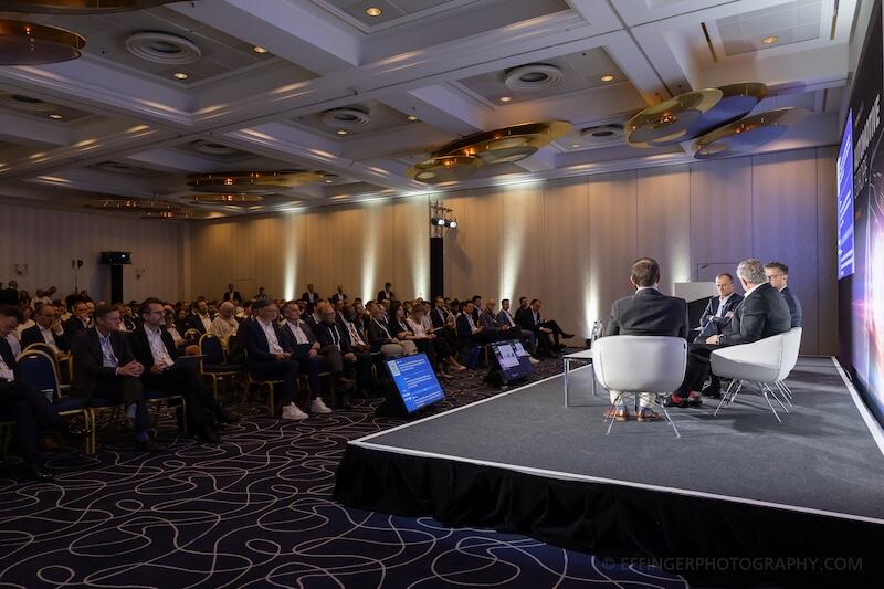 A photo of a room full of attendees with a panel on stage.