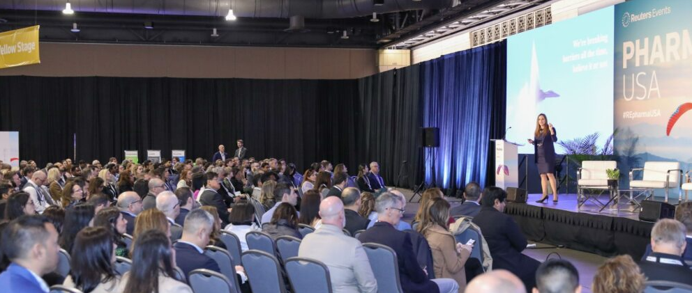 PHARMA USA conference talk; presenter on stage with large screen while a packed audience listens, "Yellow Stage" sign visible at left.
