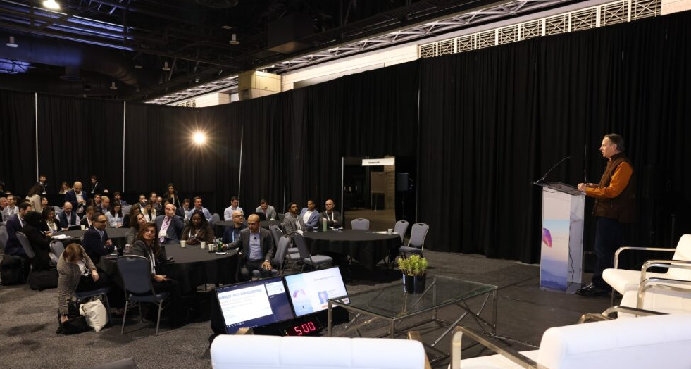 Speaker at a podium presents to a seated audience in a curtained breakout area; timer and monitors visible on stage.