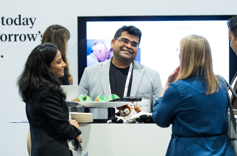 Attendees networking at an expo booth, with a smiling man in a grey blazer and lanyard conversing with two women. A plate of green-frosted donuts and a stuffed animal toy are visible on the counter in the foreground.