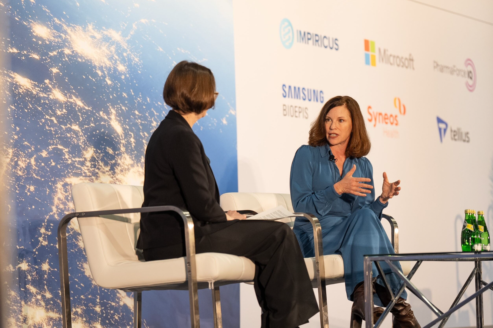 Two speakers in conversation on stage at a pharma industry conference, with sponsor logos including Impiricus, Microsoft, Samsung Bioepis, Syneos Health, and Tellius visible in the background.