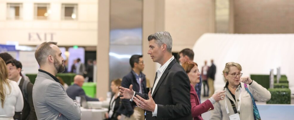 Attendees network in an open expo area; two people in foreground gesture while others converse near a white inflatable structure.