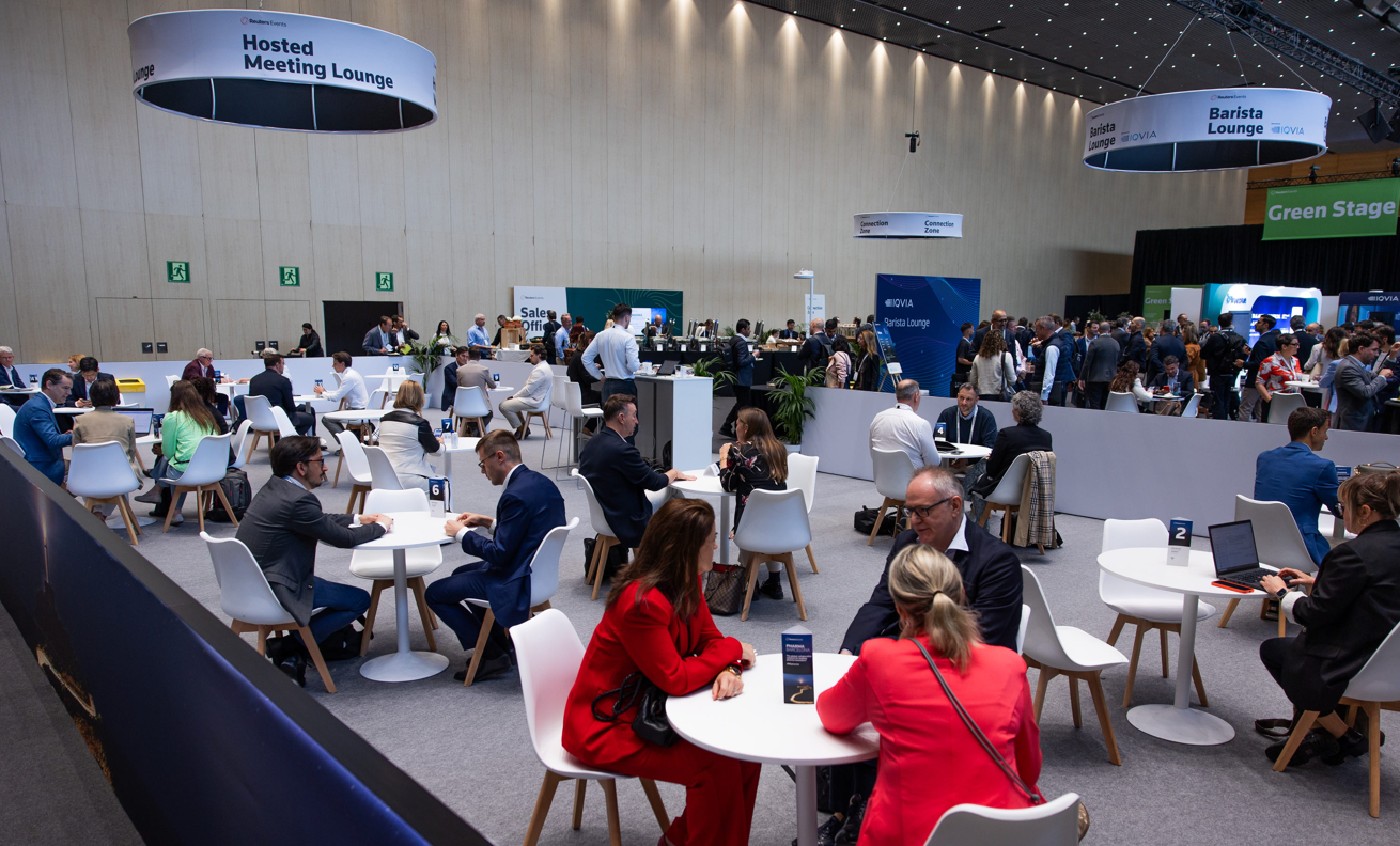 A wide overhead view of the busy Hosted Meeting Lounge and IQVIA Barista Lounge at Reuters Events Pharma Barcelona, with pharma professionals seated at numbered white round tables for one-to-one meetings, the Sales Office desk visible in the background, and the Green Stage and IQVIA exhibition booth visible to the right.