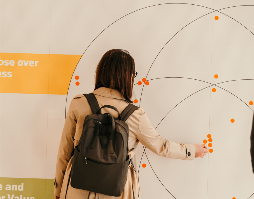 A person in a beige trench coat and backpack points to a large wall display featuring intersecting lines and orange dots at a conference booth