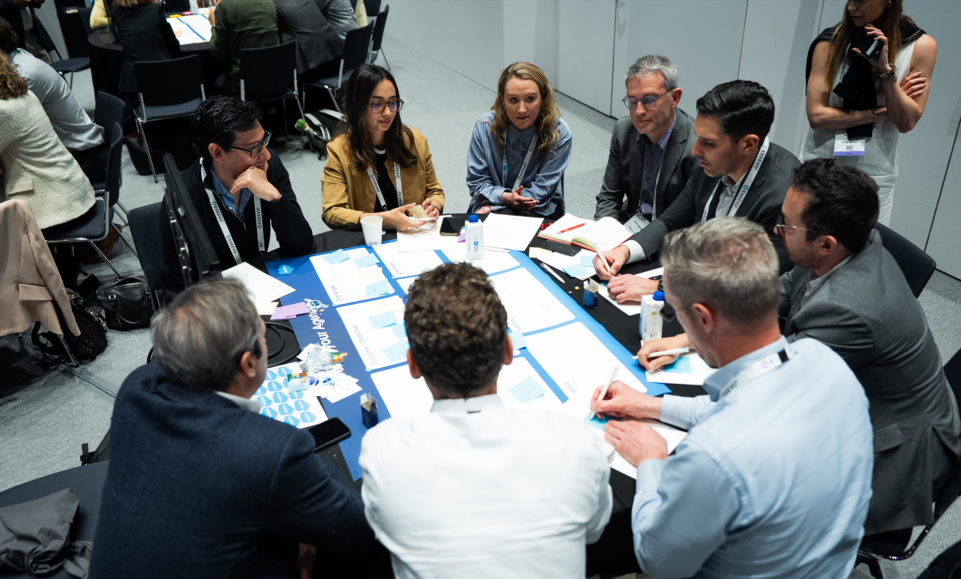A group of approximately eight pharma professionals engaged in an intensive workshop session at Reuters Events Pharma Barcelona, seated around a large table covered with printed cards, sticky notes, and worksheets, with Exeevo-branded lanyards visible and additional delegates working at tables in the background.