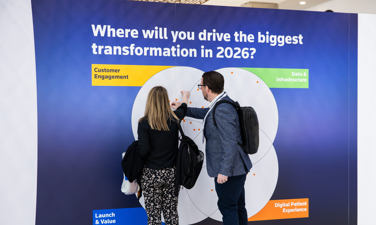 Two Reuters Events Pharma Barcelona attendees — a woman and a man wearing a conference lanyard and backpack — placing orange dot stickers on a large interactive wall display titled "Where will you drive the biggest transformation in 2026?" featuring four categories: Customer Engagement, Data & Infrastructure, Digital Patient Experience, and Launch & Value.