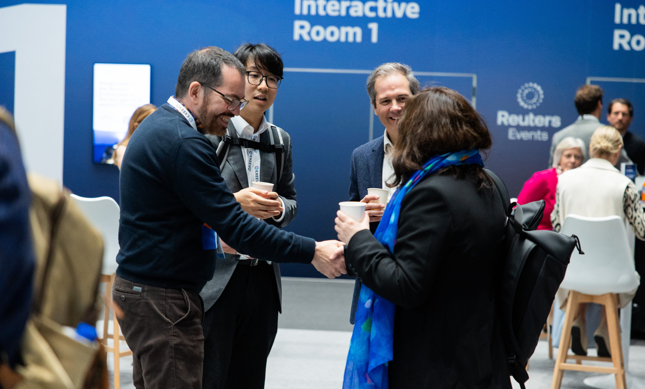 Four pharma professionals networking with coffee cups outside Interactive Room 1 at Reuters Events Pharma Barcelona, with one attendee shaking hands warmly, Exeevo-branded lanyards visible, and the Reuters Events logo on the blue wall behind them.