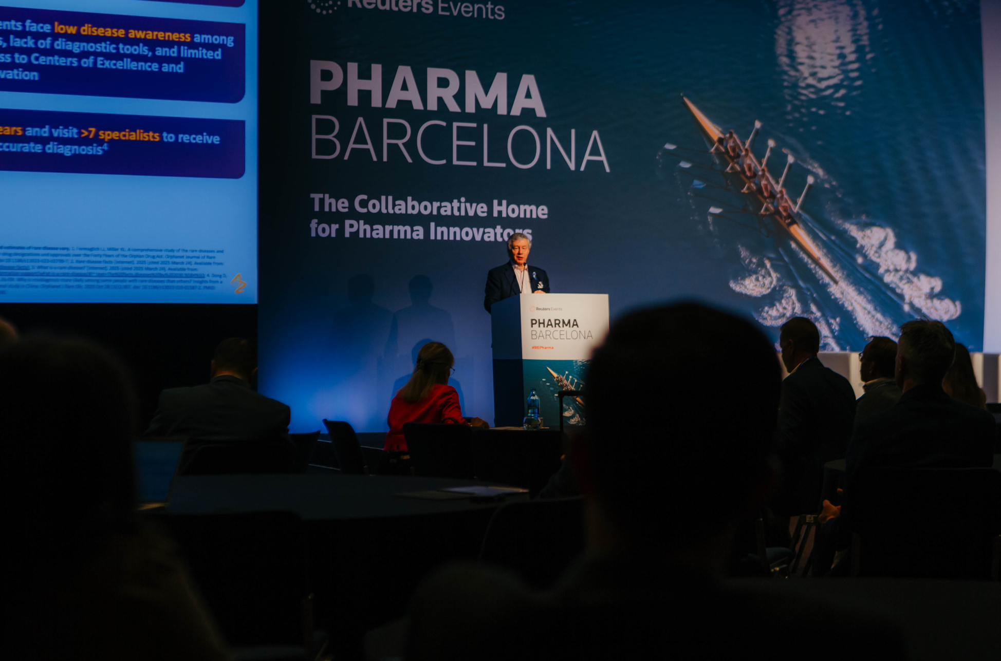 A speaker stands at a podium in front of a large screen displaying "PHARMA BARCELONA" and an image of a rowing team. Attendees are seated in the foreground, facing the speaker.