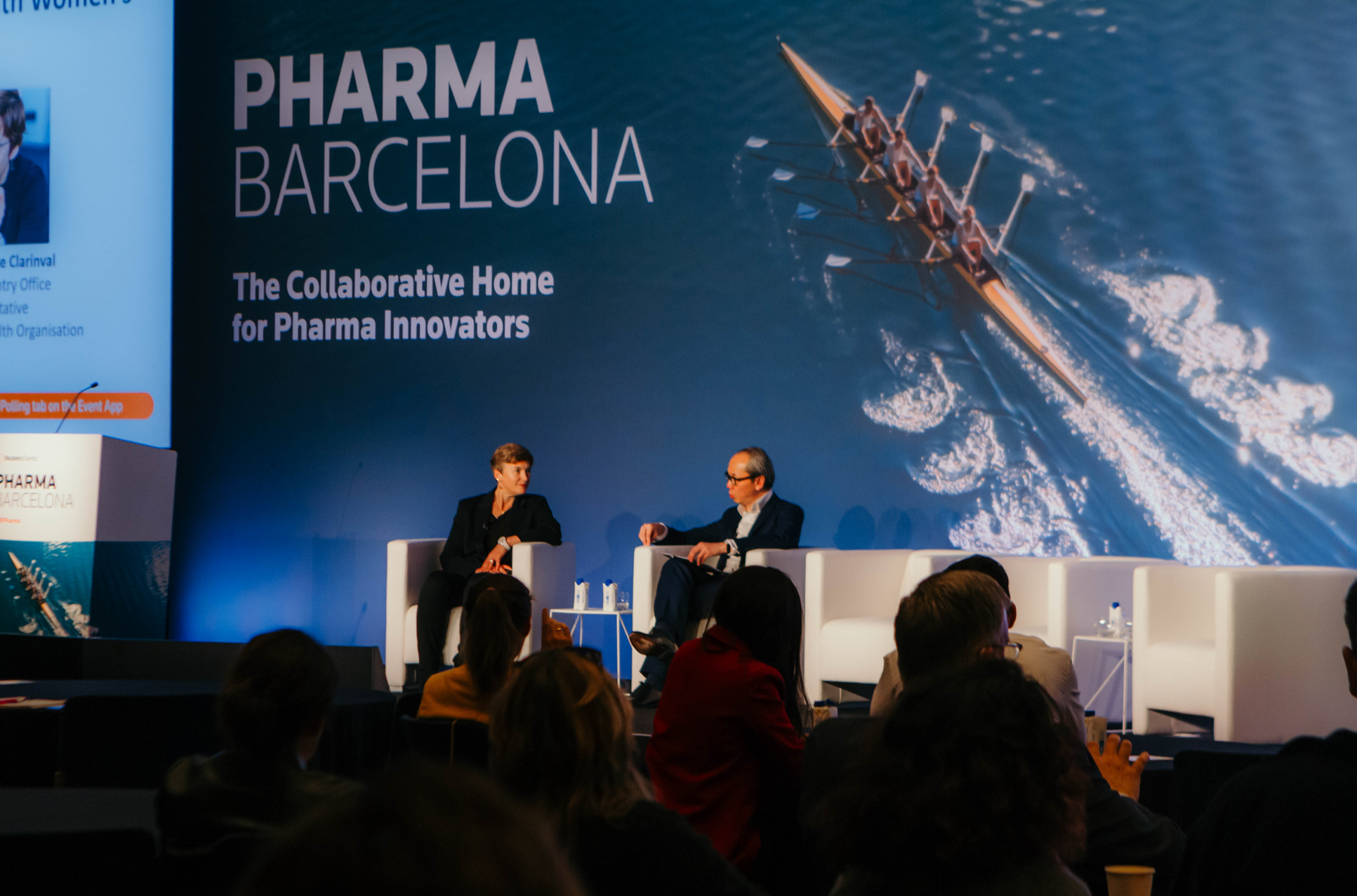 Two panelists sit on white armchairs on a stage, facing each other and smiling, with an audience in the foreground. Behind them, a large screen displays "PHARMA BARCELONA: The Collaborative Home for Pharma Innovators" next to an image of a rowing crew.