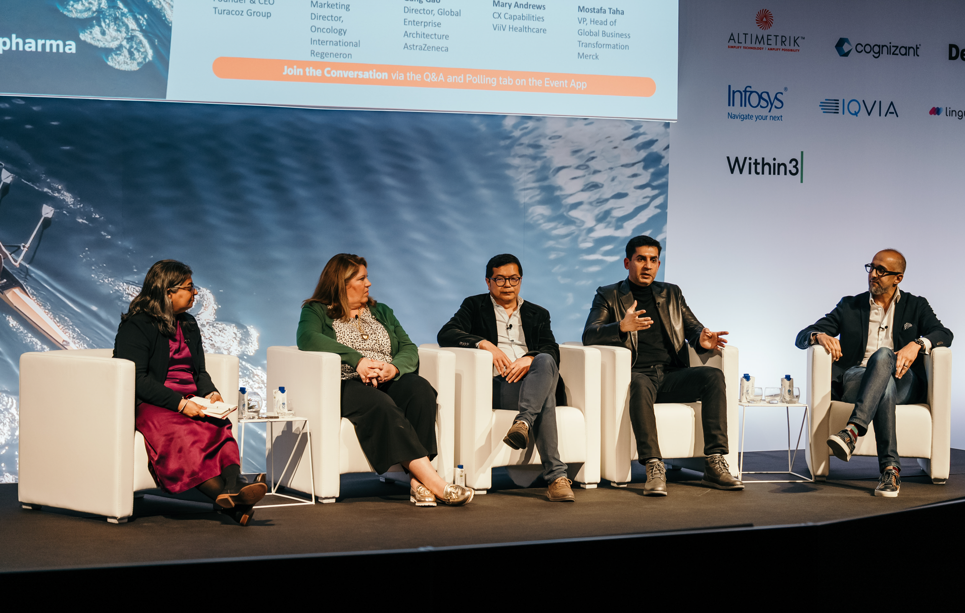 A panel of five speakers sit in white armchairs on a stage during a conference. The speaker on the far right gestures with his hands while speaking. A large screen behind them displays "PHARMA BARCELONA" with logos of sponsors and names of other speakers and companies.
