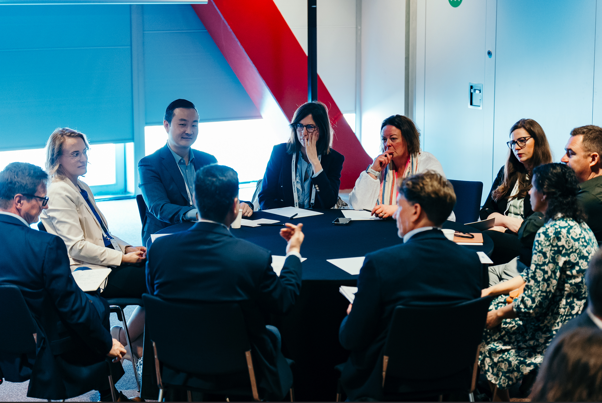 A group of people seated around a dark table are engaged in a discussion during a conference.