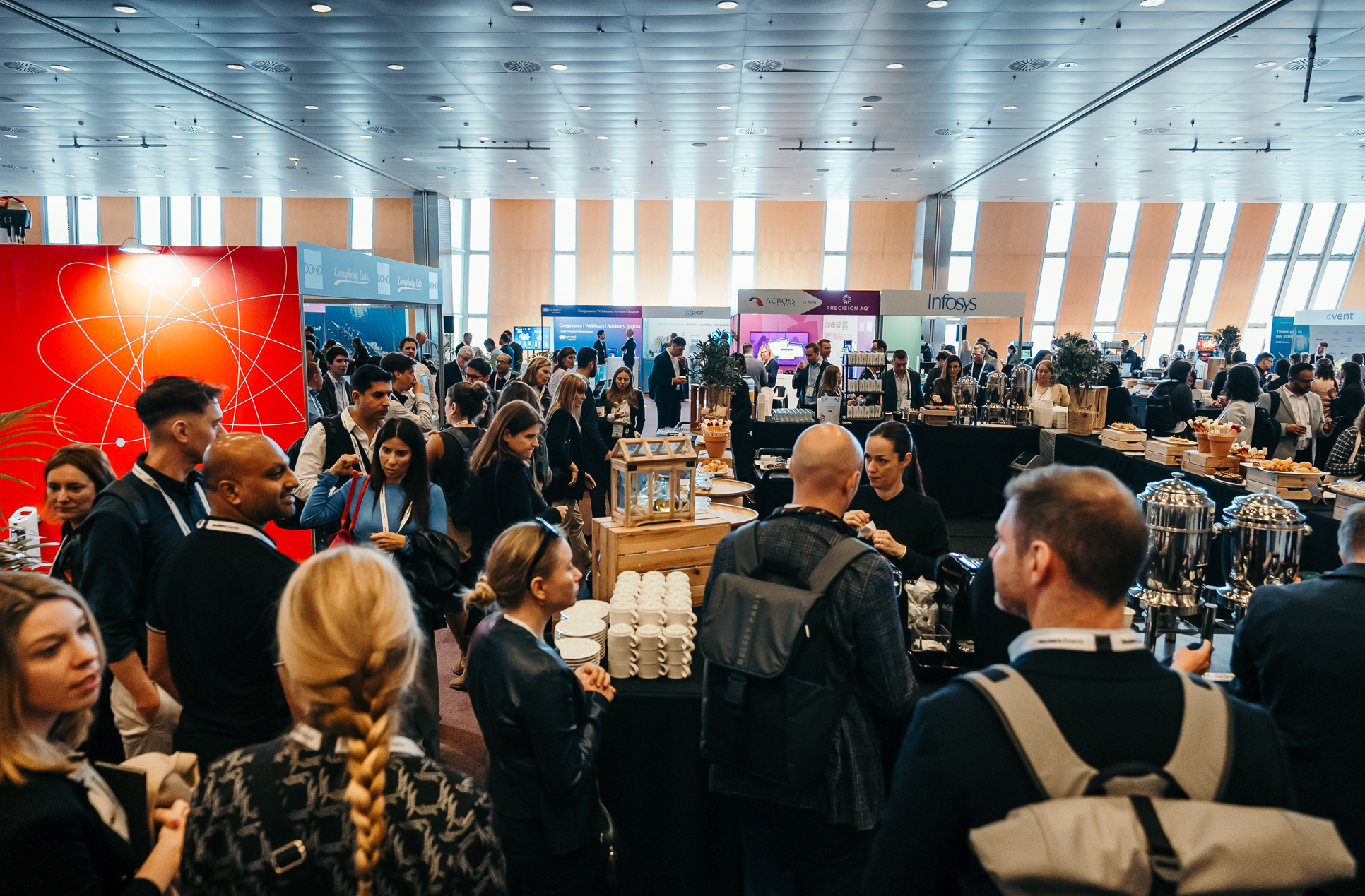 A large number of people gathered in a spacious conference hall, with various exhibition booths and catering stations visible.