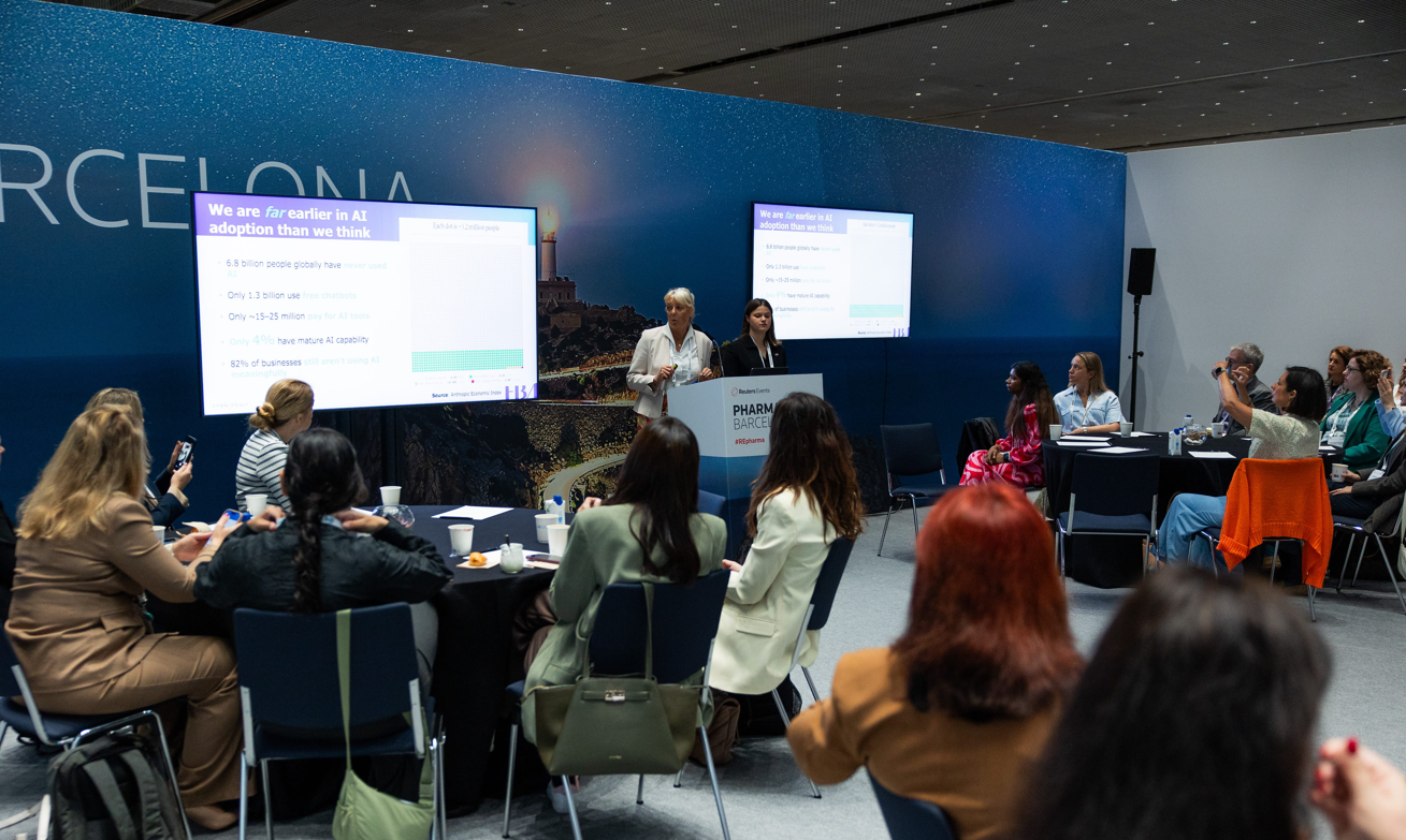 Two female speakers present at the Reuters Events Pharma Barcelona podium to a room of predominantly female delegates seated at round tables, with two large screens displaying a slide titled "We are far earlier in AI adoption than we think," sourced from the McKinsey Economic Index, and the Pharma Barcelona branded backdrop visible behind them.