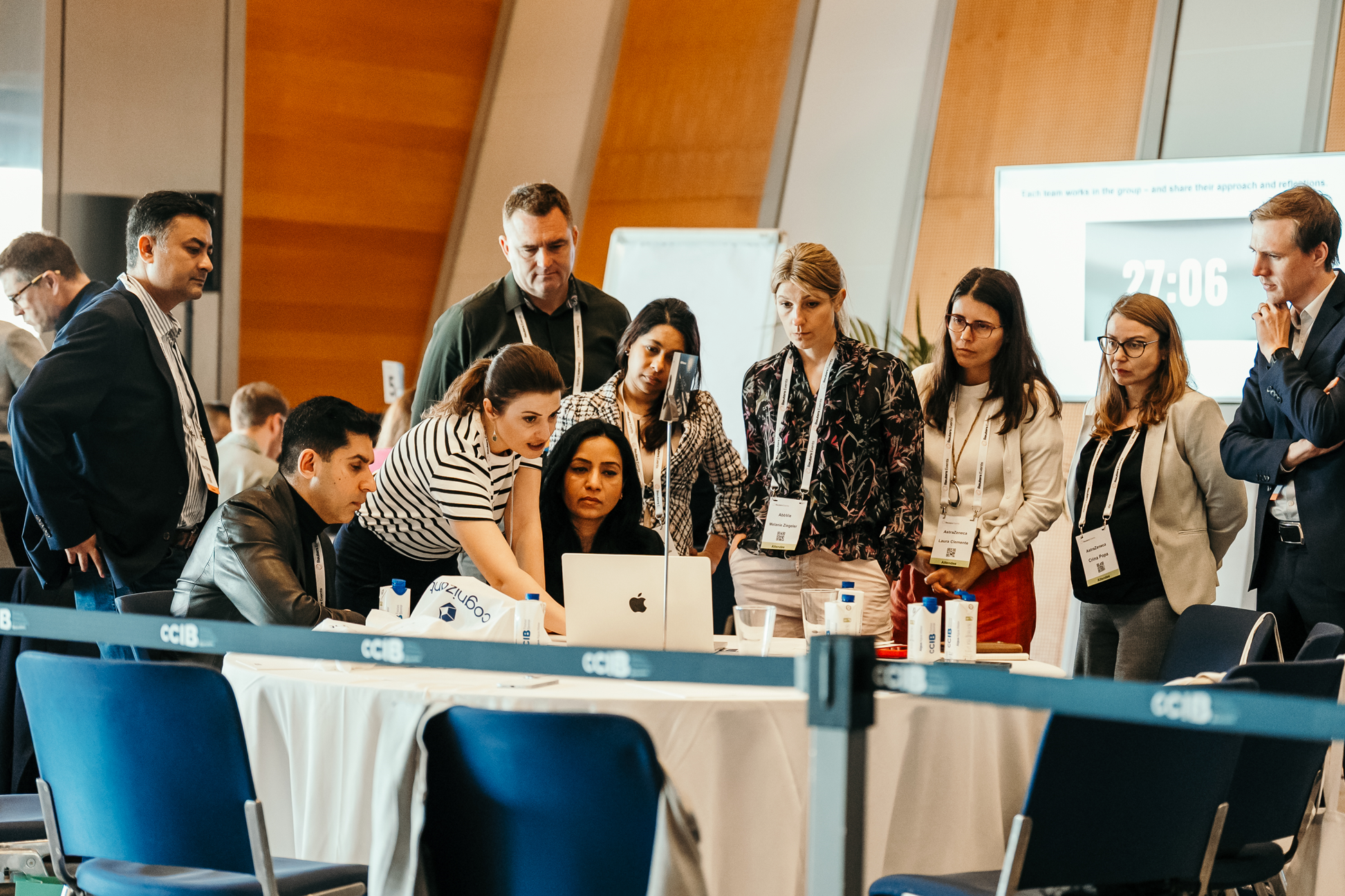 A group of professionals, some seated and some standing, are intently gathered around a laptop on a table during a conference. In the background, a screen displays "27:06."