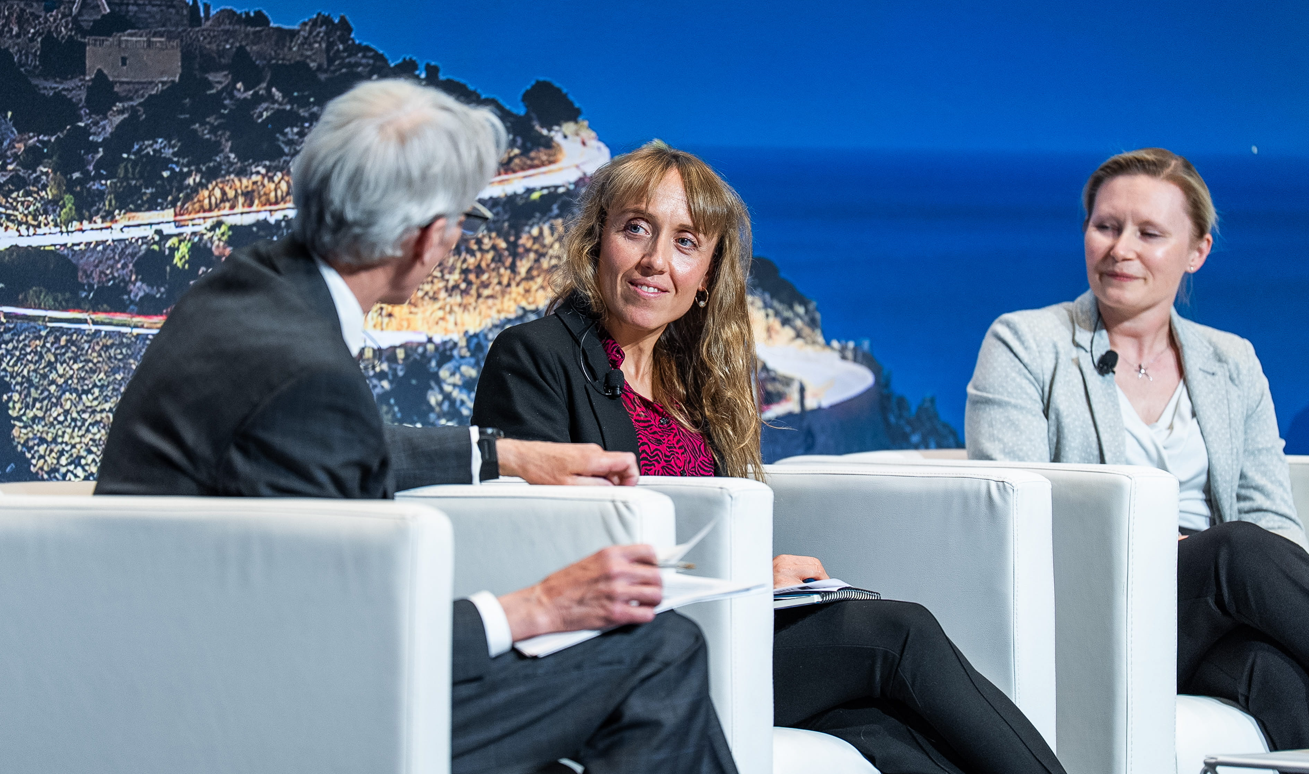 A close-up of three panellists in a fireside chat on the main stage at Reuters Events Pharma Barcelona, with a male moderator with grey hair facing two female speakers seated in white armchairs, all wearing lapel microphones, against a dramatic coastal cliff and lighthouse backdrop.