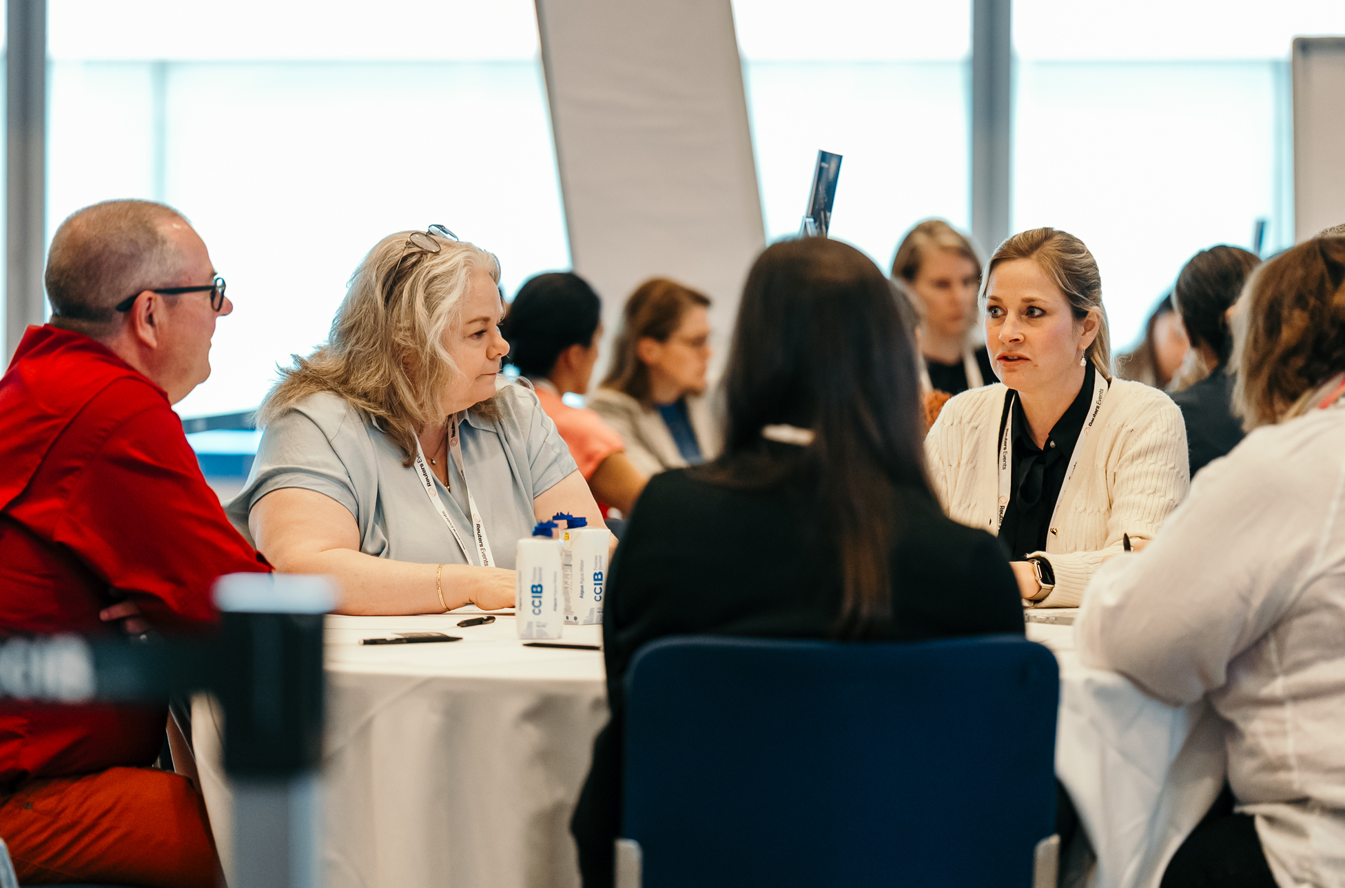 A group of professionals are seated around a white table, actively engaged in a discussion during a conference. The focus is on their interaction and participation.