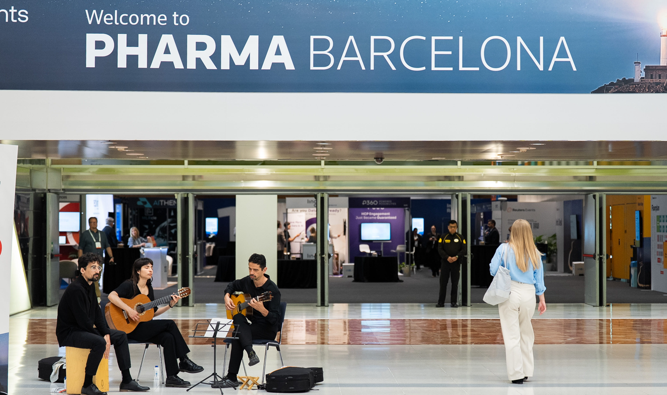 A flamenco-style trio of musicians dressed in black — two guitarists and a percussionist — perform in the entrance foyer of Reuters Events Pharma Barcelona, with a large "Welcome to Pharma Barcelona" banner overhead, exhibition booths visible through glass doors in the background, and a female conference attendee watching the performance.