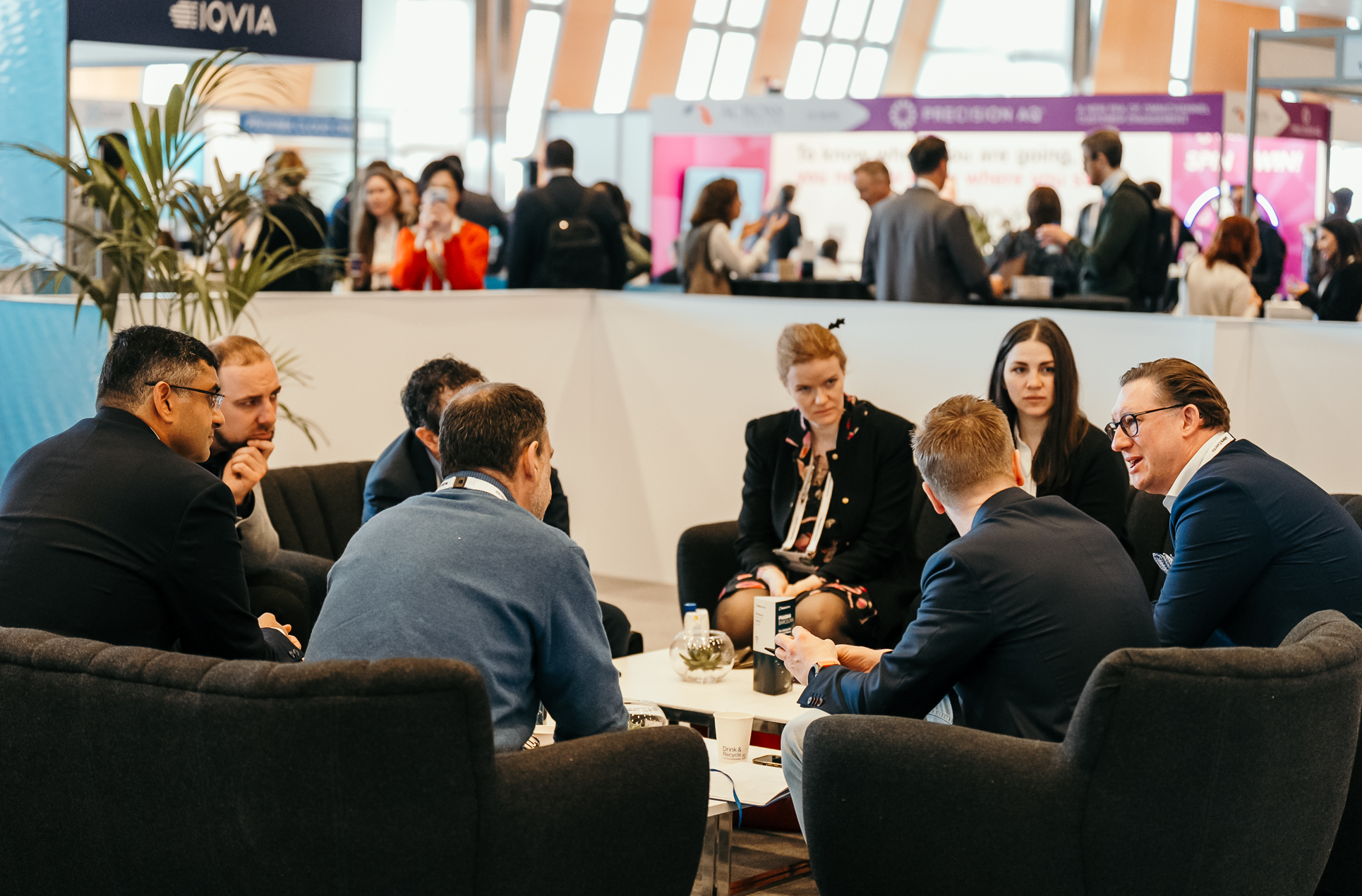 Attendees at a pharma conference sit in a circle, engaged in discussion. Several people are visible in the background networking at various booths.