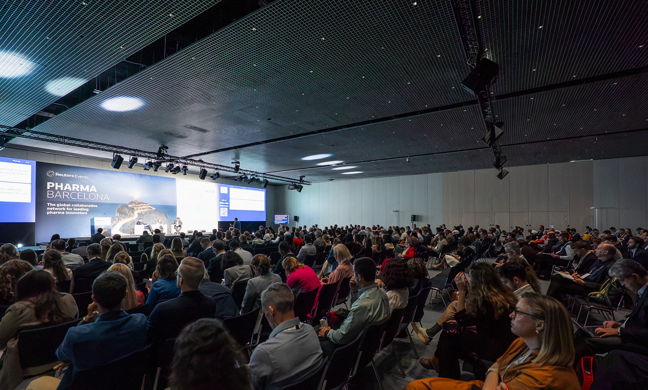 A wide-angle view of a packed plenary session at Reuters Events Pharma Barcelona, with hundreds of pharma professionals seated in rows of chairs facing the main stage, which displays the Pharma Barcelona branding and the tagline "The global collaborative network for leading pharma innovators" on large LED screens, with professional stage lighting overhead.