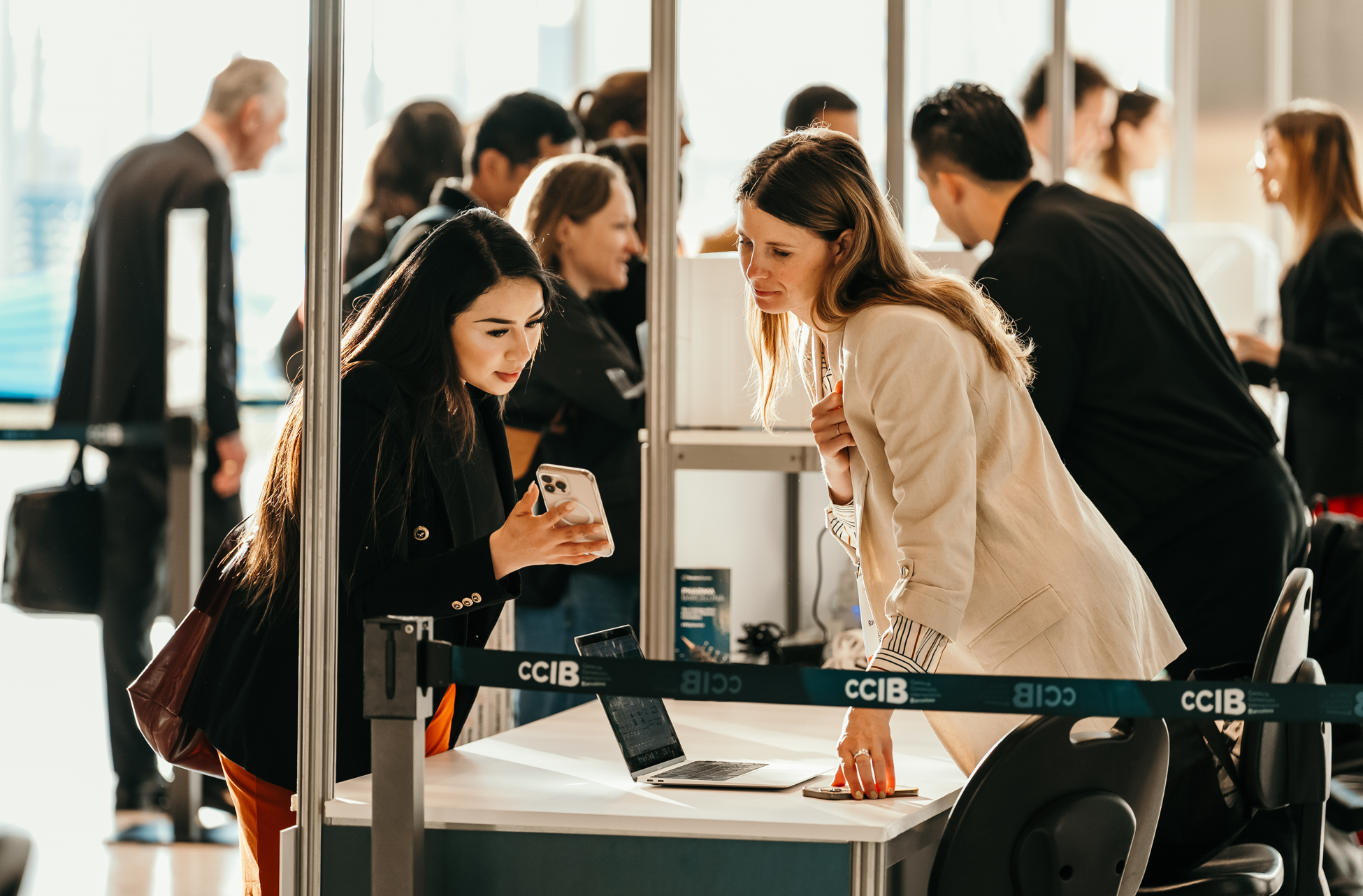 Two women are interacting at a registration desk during a conference. One woman is looking at her phone, while the other leans in, observing. A laptop is open on the desk, and a banner with "CCIB" is visible in the foreground.