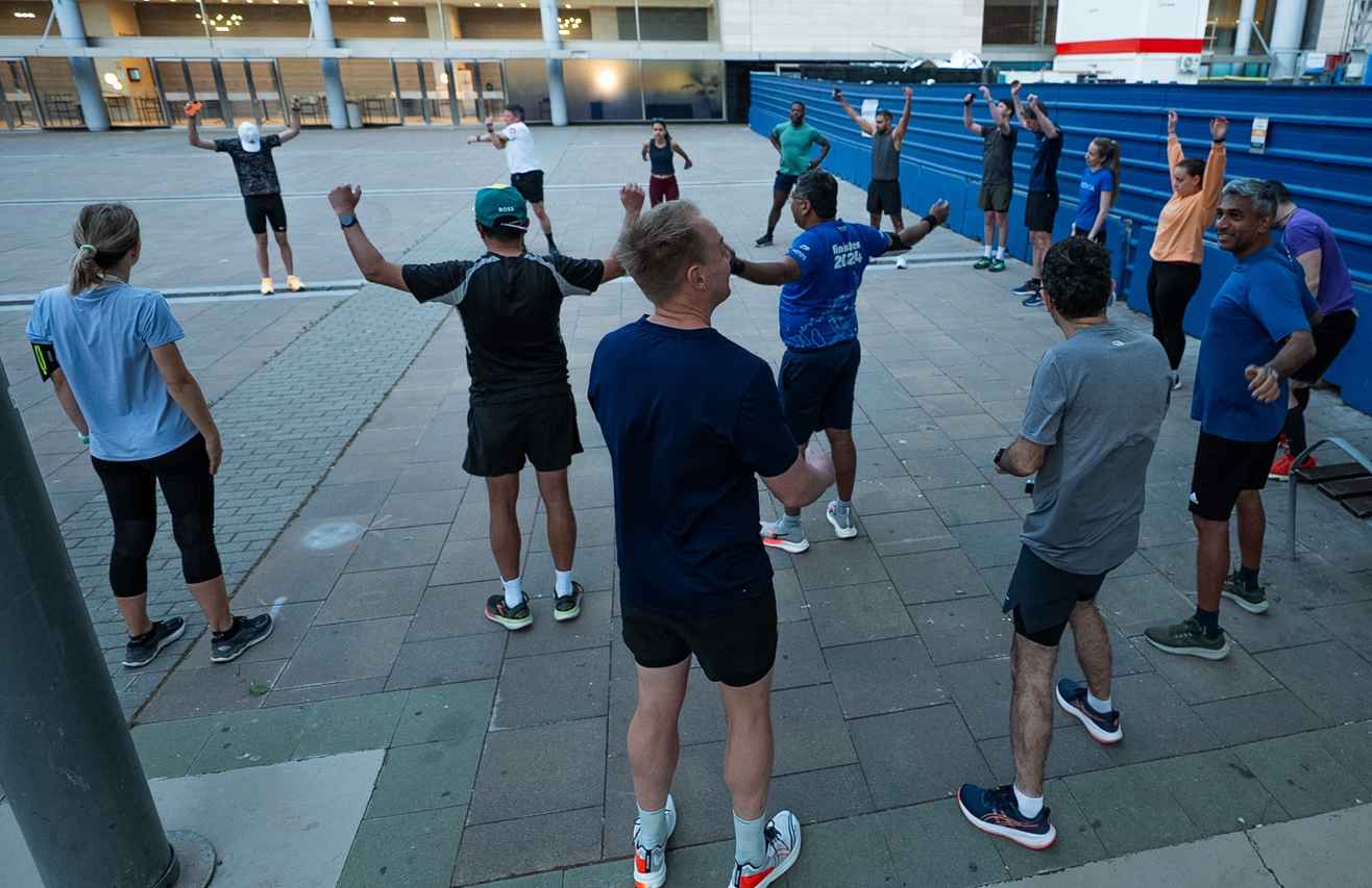 A group of approximately fifteen Reuters Events Pharma Barcelona conference attendees in athletic wear performing warm-up stretches in an outdoor plaza at dawn, preparing for a morning group run, with the conference venue building visible in the background.