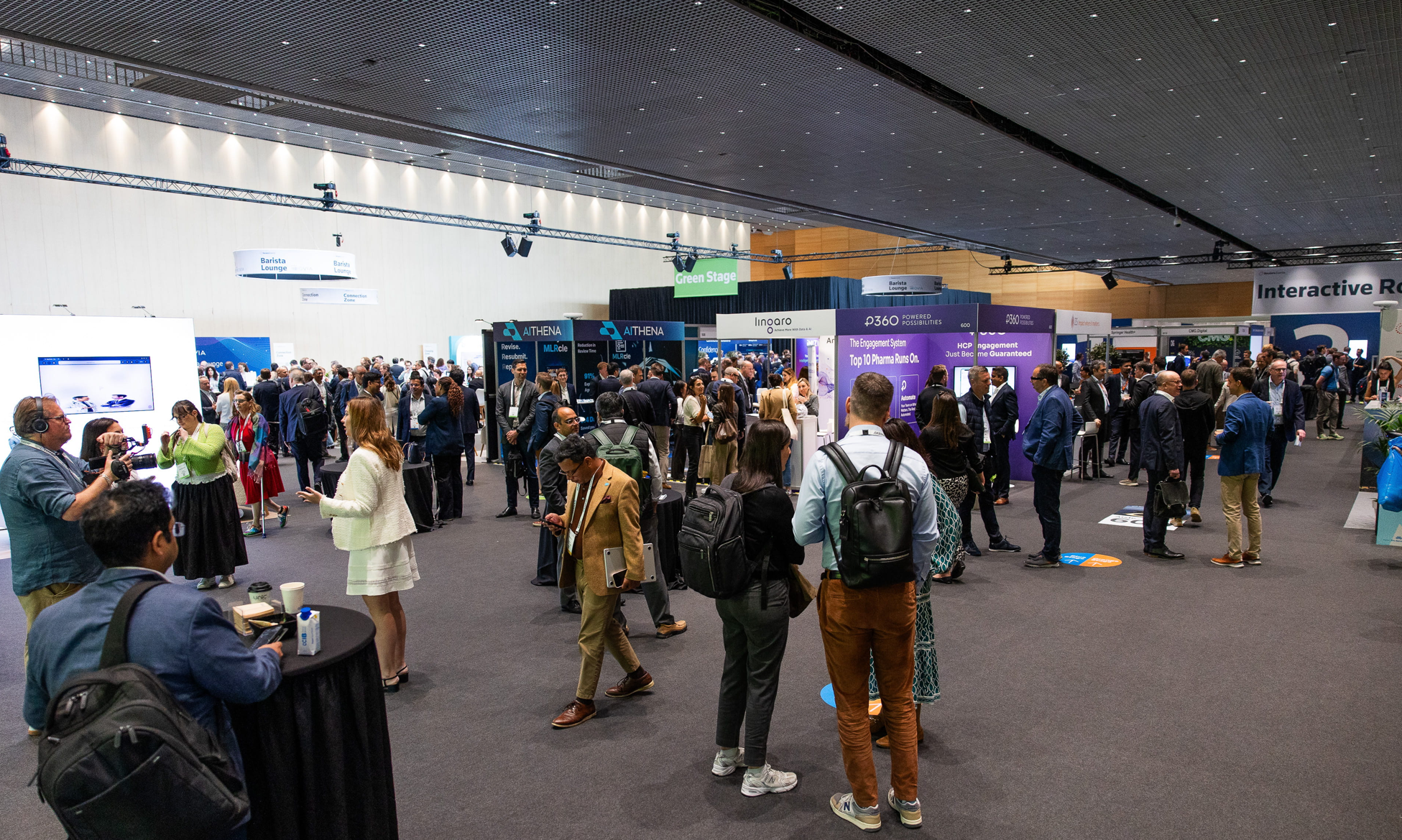 A wide-angle view of the busy exhibition hall at Reuters Events Pharma Barcelona, with hundreds of pharma professionals browsing sponsor booths including AITHENA, Linoaro, P360, and others, the Green Stage visible in the centre background, and the Interactive Rooms signage visible to the right.