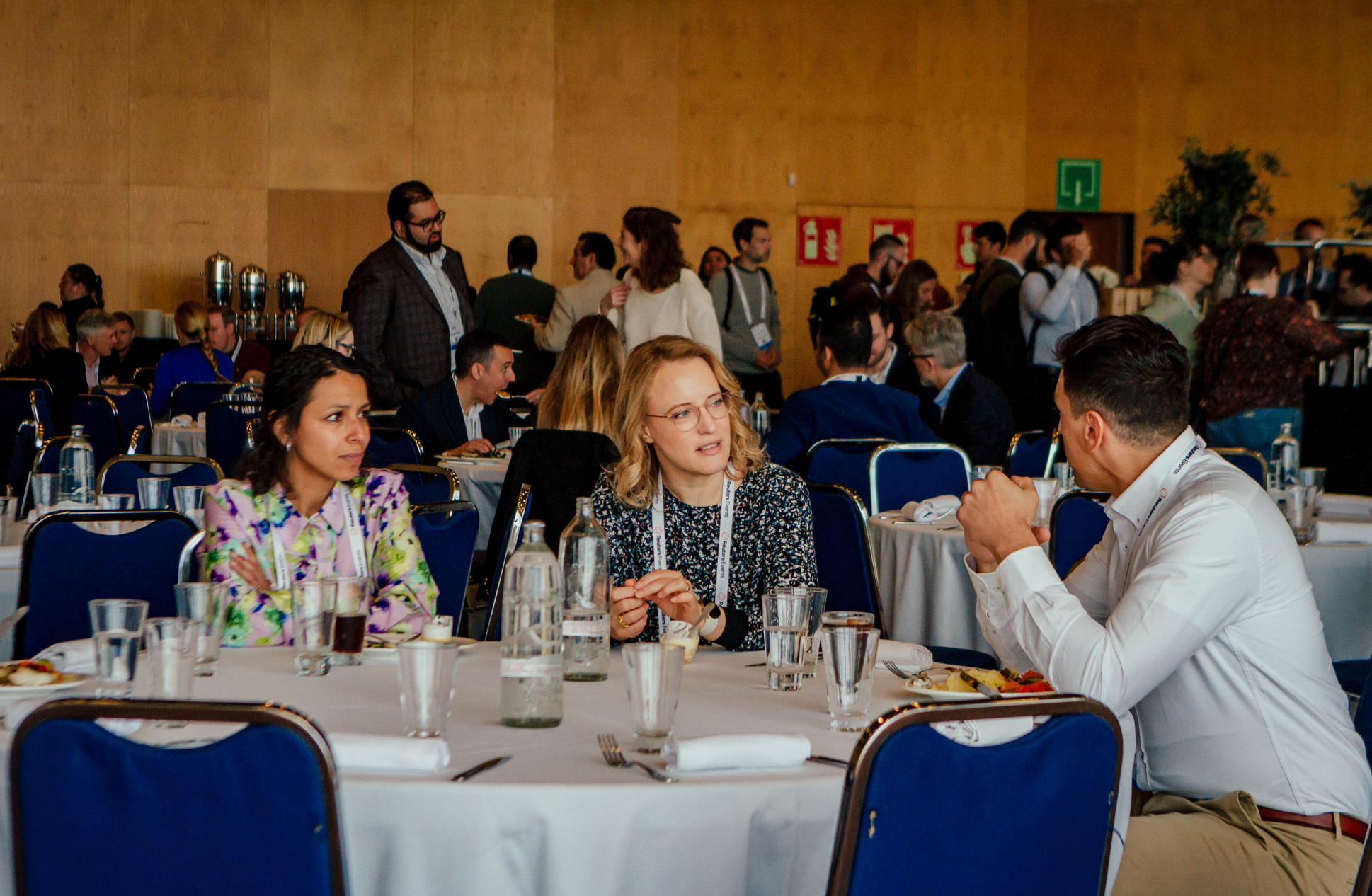 Three people seated at a round table, engaged in conversation, with other attendees and a buffet line visible in the background at a conference.