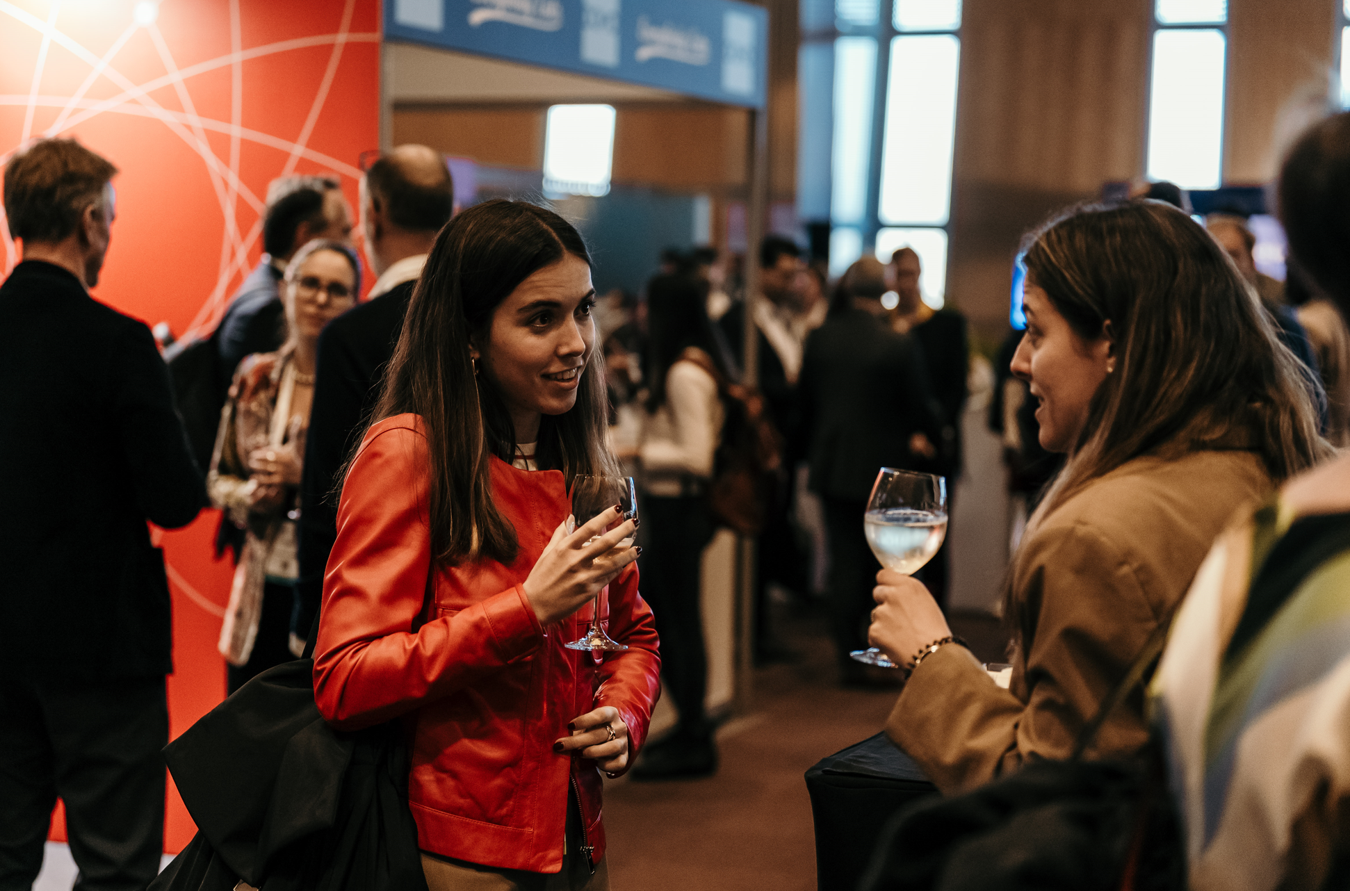 Two women engage in conversation, holding glasses of wine, amidst a crowded networking event at a conference.