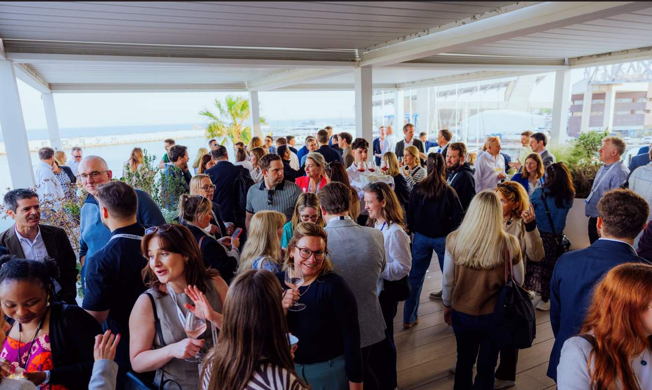A large group of pharma industry professionals networking and socialising with drinks at an outdoor waterfront terrace event during Reuters Events Pharma Barcelona, with palm trees, moored yachts, and the Barcelona marina visible in the background under a clear sky.
