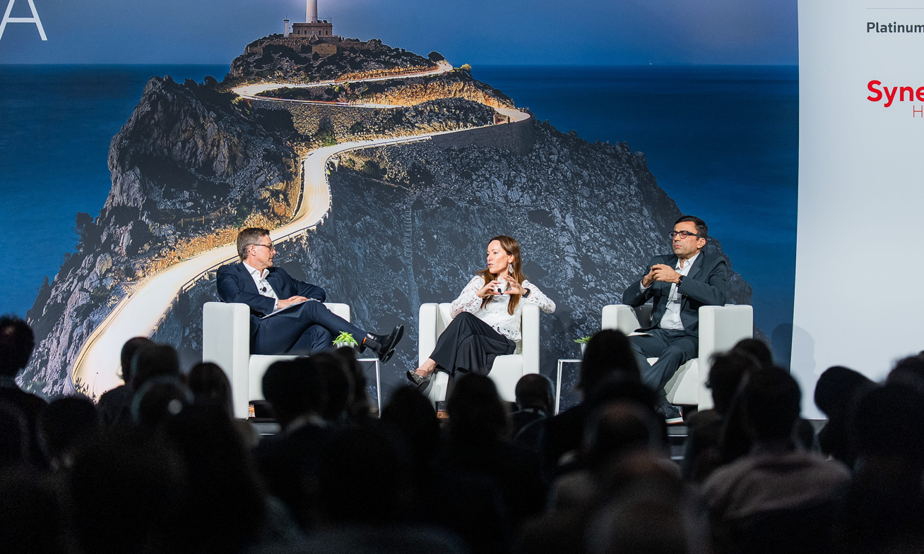 Three pharma industry leaders — two men and a woman — engaged in a panel discussion on white armchair seats on the main stage at Reuters Events Pharma Barcelona, with a dramatic lighthouse and coastal road backdrop projected on the large screen behind them and a Syneos Health Platinum Sponsor banner visible to the right, with a silhouetted audience in the foreground.