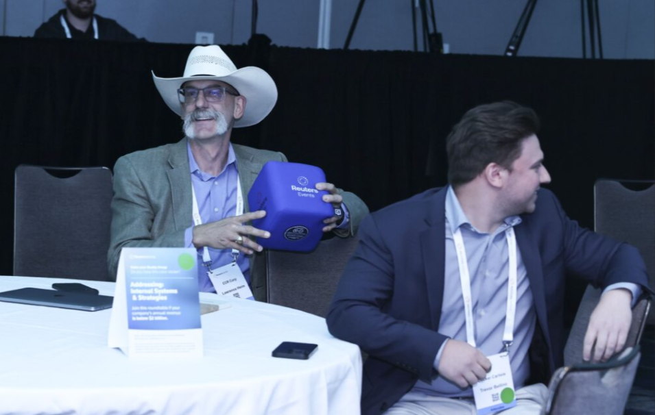 Attendees at Responsible Business USA 2025 seated at a roundtable; man in a cowboy hat holds a Reuters Events throwable mic cube as another looks on.